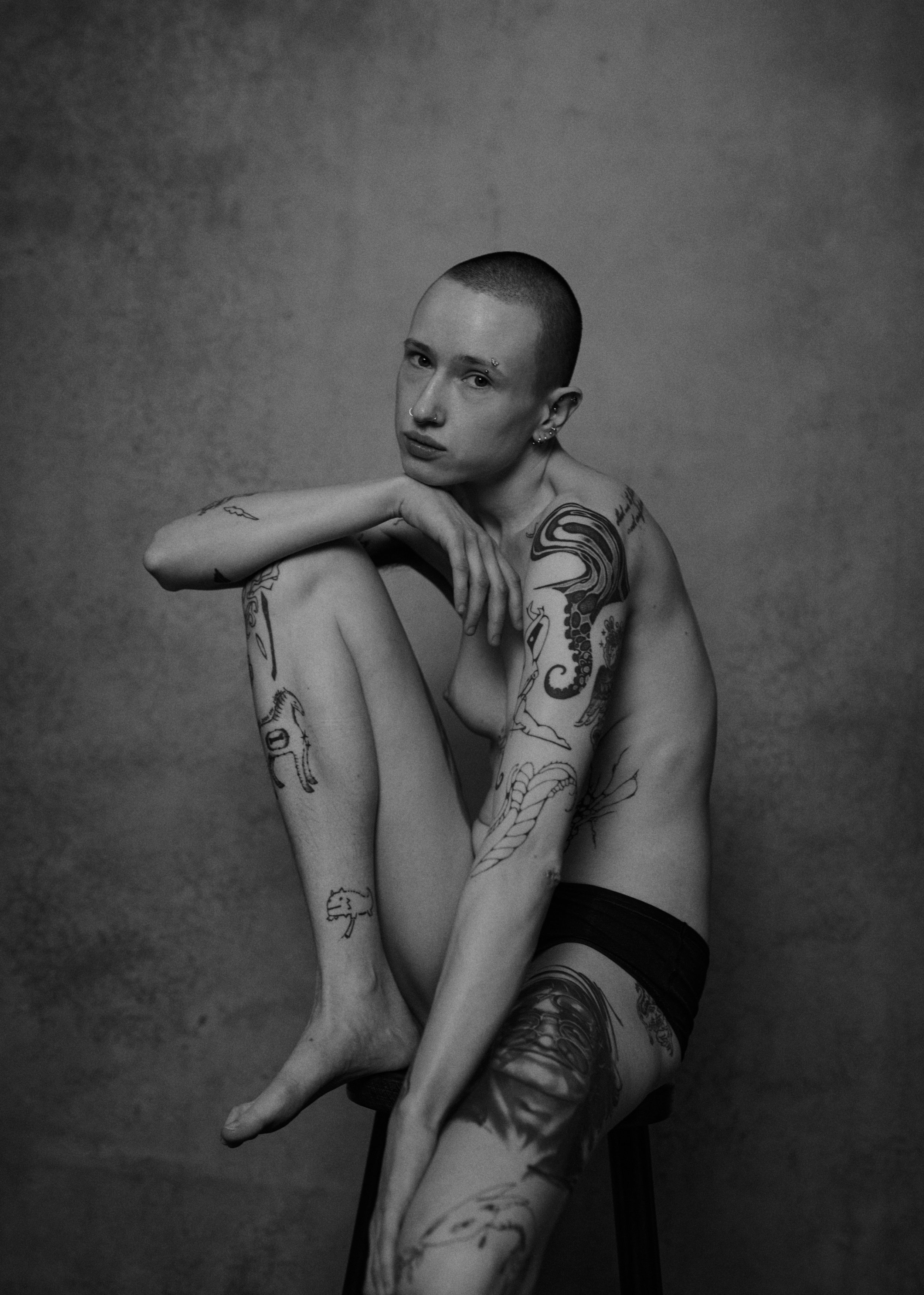 Schwarz-weiß-Fotografie eines nackten Menschen mit Tattoos, der auf einem Stuhl sitzt, das Bein um die Schulter gelegt, Blick direkt in die Kamera, gegen eine einfache Hintergrundwand.