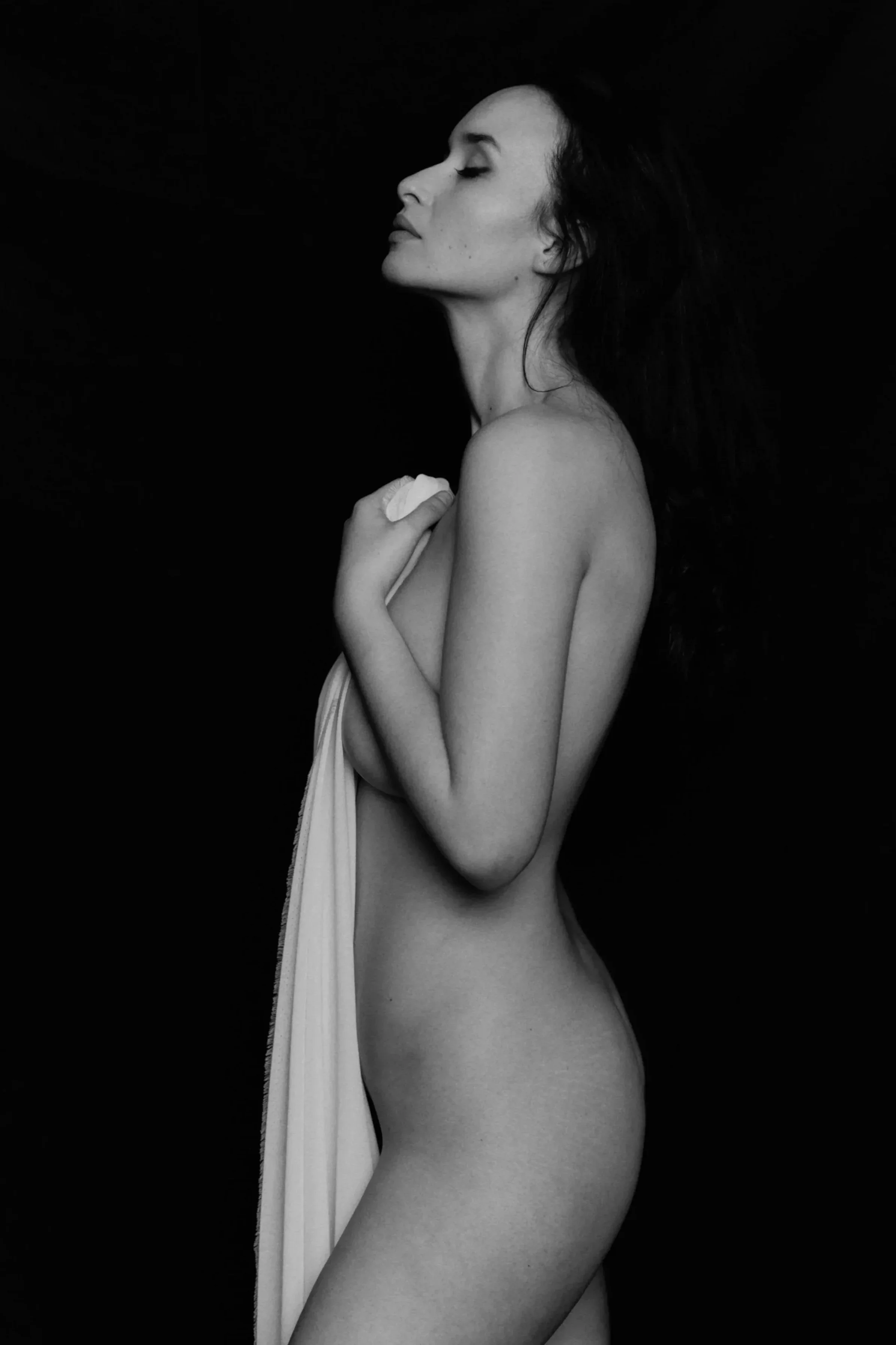 Schwarz-Weiß-Fotografie einer Frau, die nackt ist, mit geschlossenen Augen, vor einem dunklen Hintergrund, die eine Stoffbahn hält.