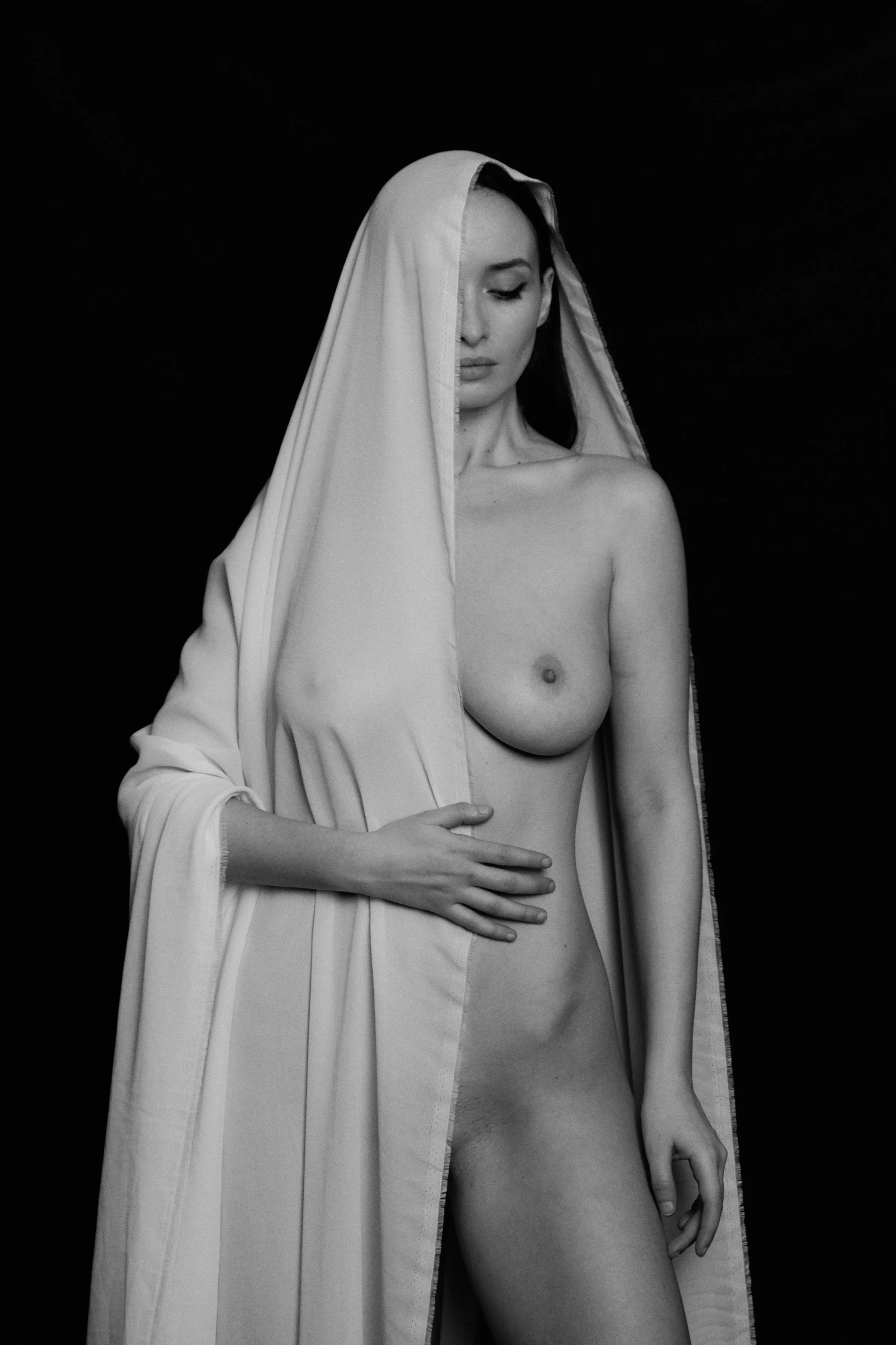 Schwarz-Weiß-Fotografie einer nackten Frau mit einem weißen Tuch über dem Kopf und der Schulter gegen einen schwarzen Hintergrund.