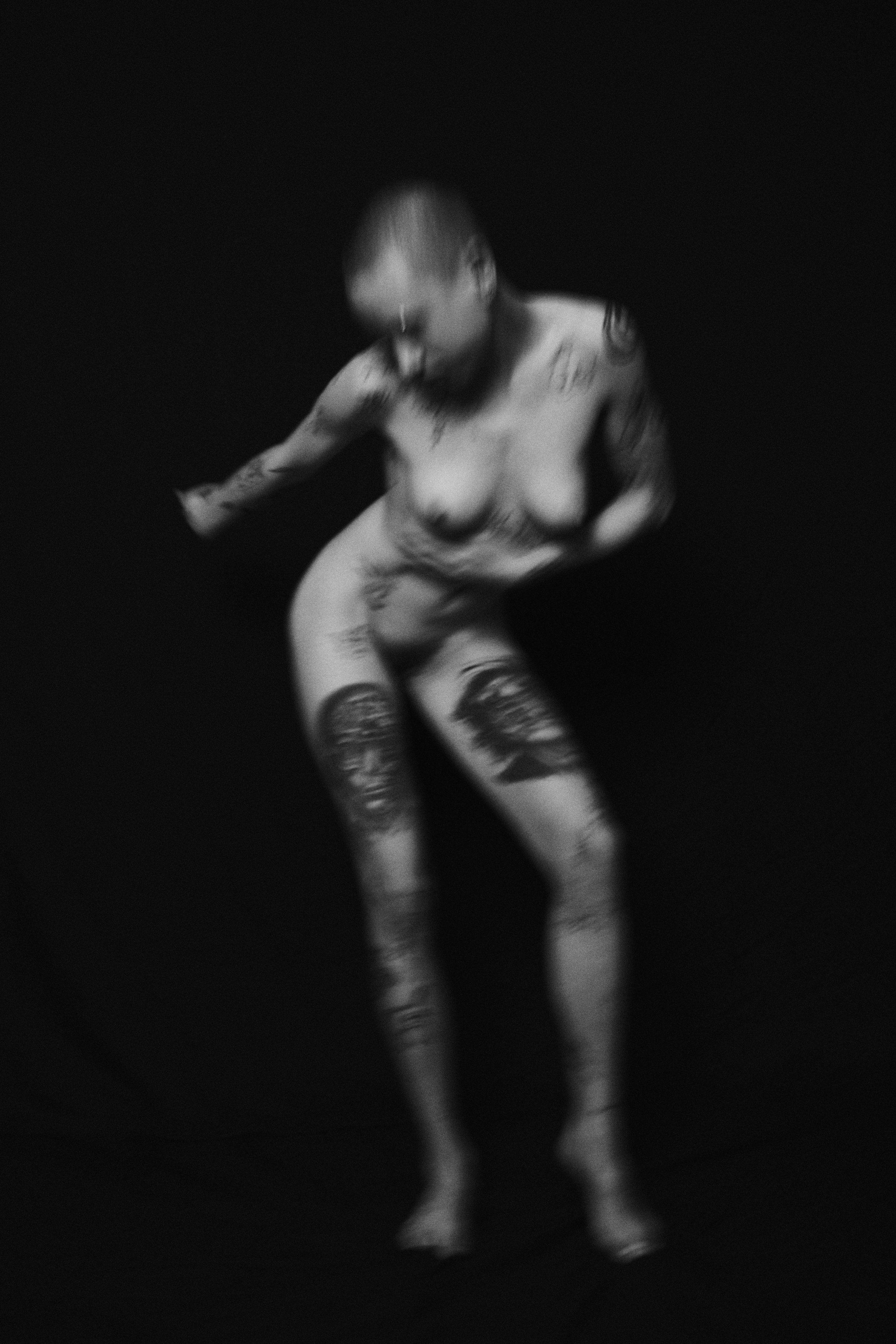 Schwarz-weiß-Foto einer nackt tätowierten Frau, die auf einem schwarzen Hintergrund steht, leicht nach vorne gebeugt ist und den Kopf gesenkt hat. Nude Art