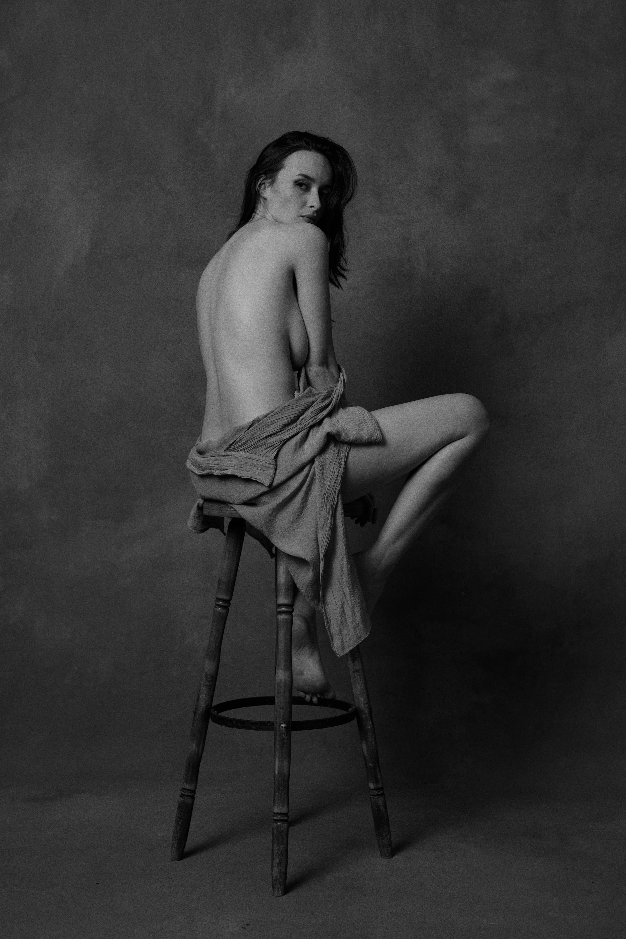 Schwarz-Weiß-Fotografie einer nackten Frau, die auf einem Holzstuhl sitzt, mit einem Tuch um die Hüften. Sie blickt nach hinten über ihre Schulter, vor einem dunklen Hintergrund. Nude Art