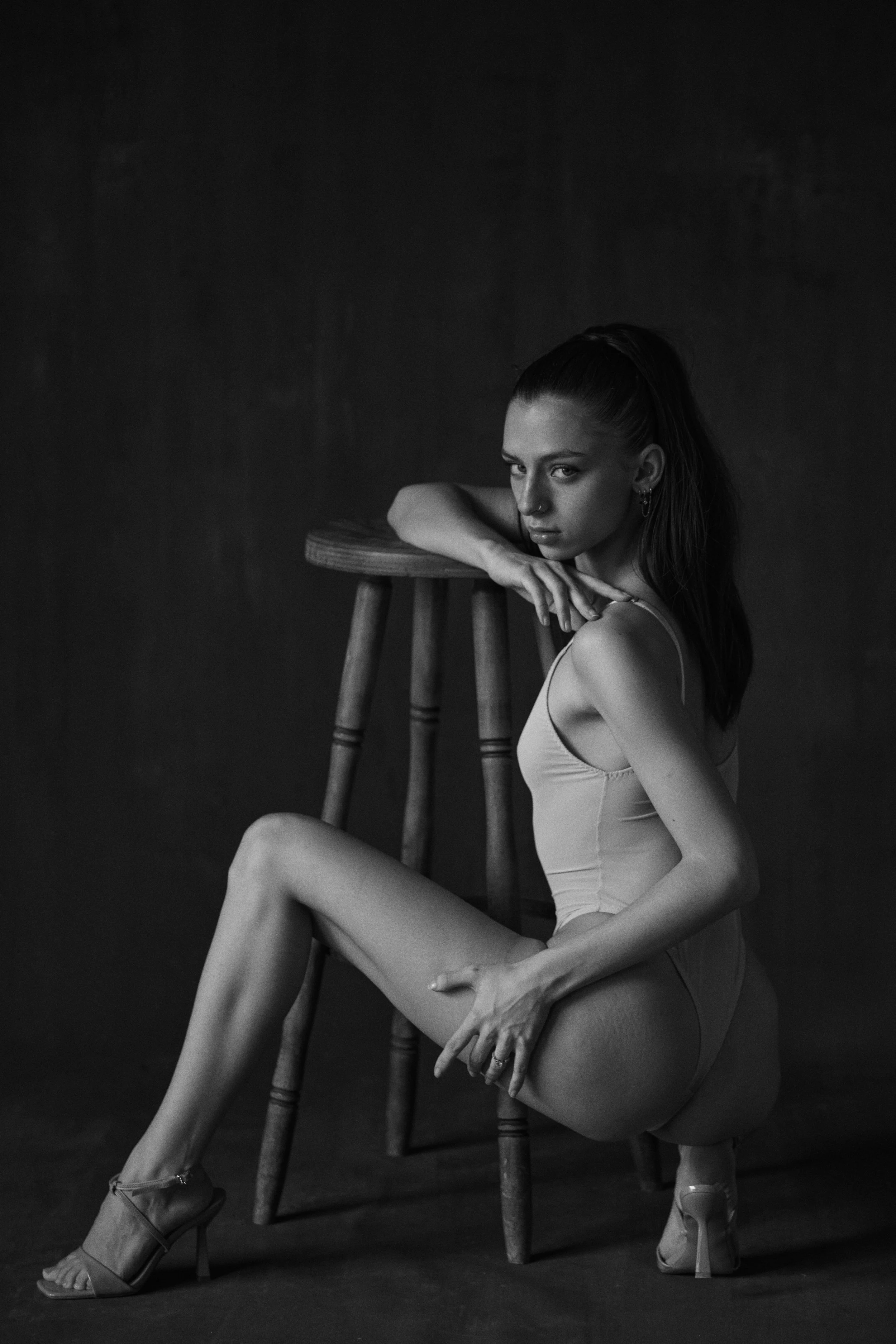 Schwarz-Weiß-Fotografie einer jungen Frau in Unterwäsche, die auf dem Boden neben einem Holzstuhl sitzt, mit nachdenklichem Blick.