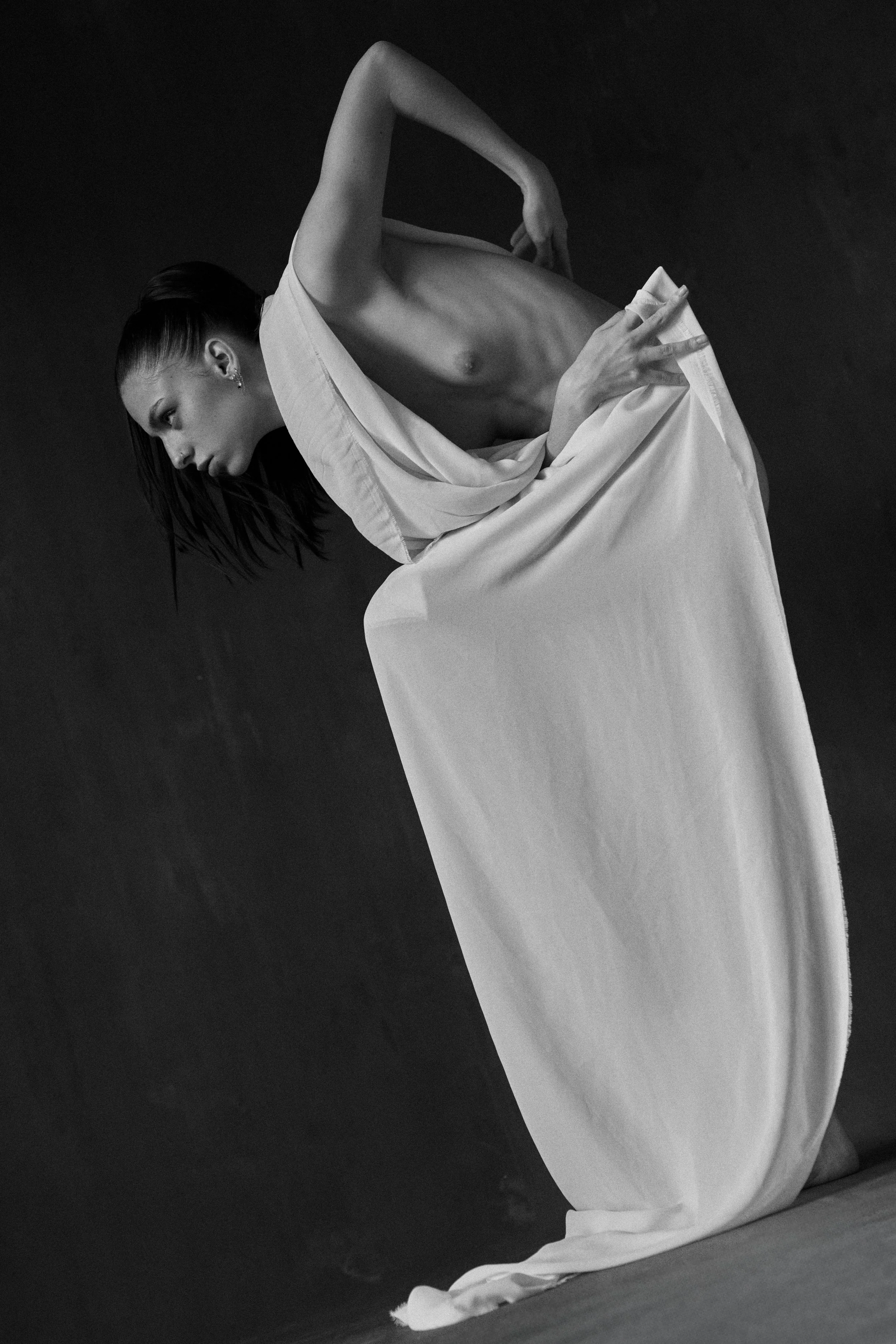 Schwarz-Weiß-Fotografie einer jungen Frau in einem langen weißen Kleid, die auf dem Boden kniet, mit ihrem Oberkörper nach vorne gebeugt, dunkler Hintergrund.