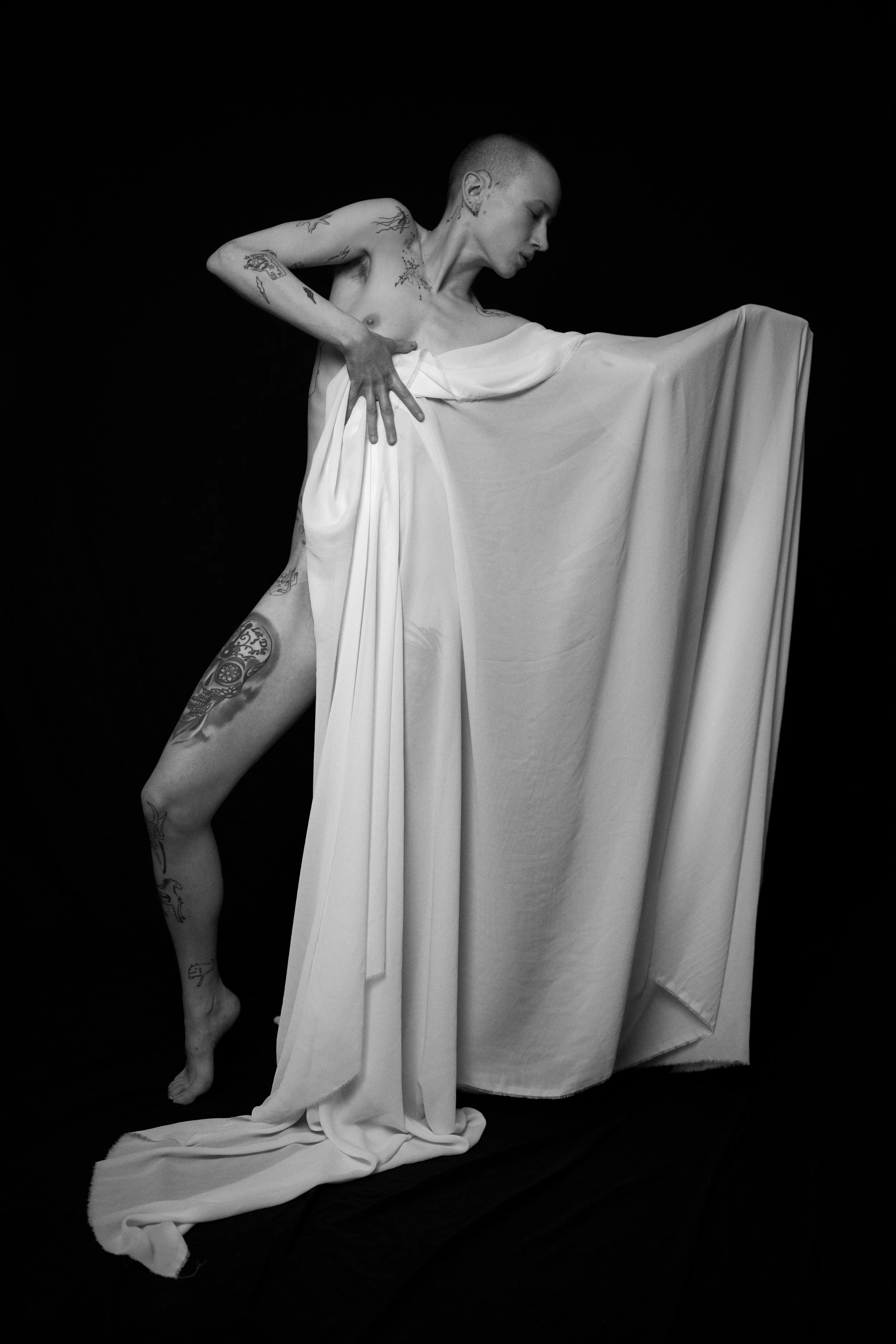Schwarz-weiß-Fotografie einer tätowierten Person, die ein weises Kleid trägt und es auf eine dramatische Weise hochhält, vor schwarzem Hintergrund. Nude Art