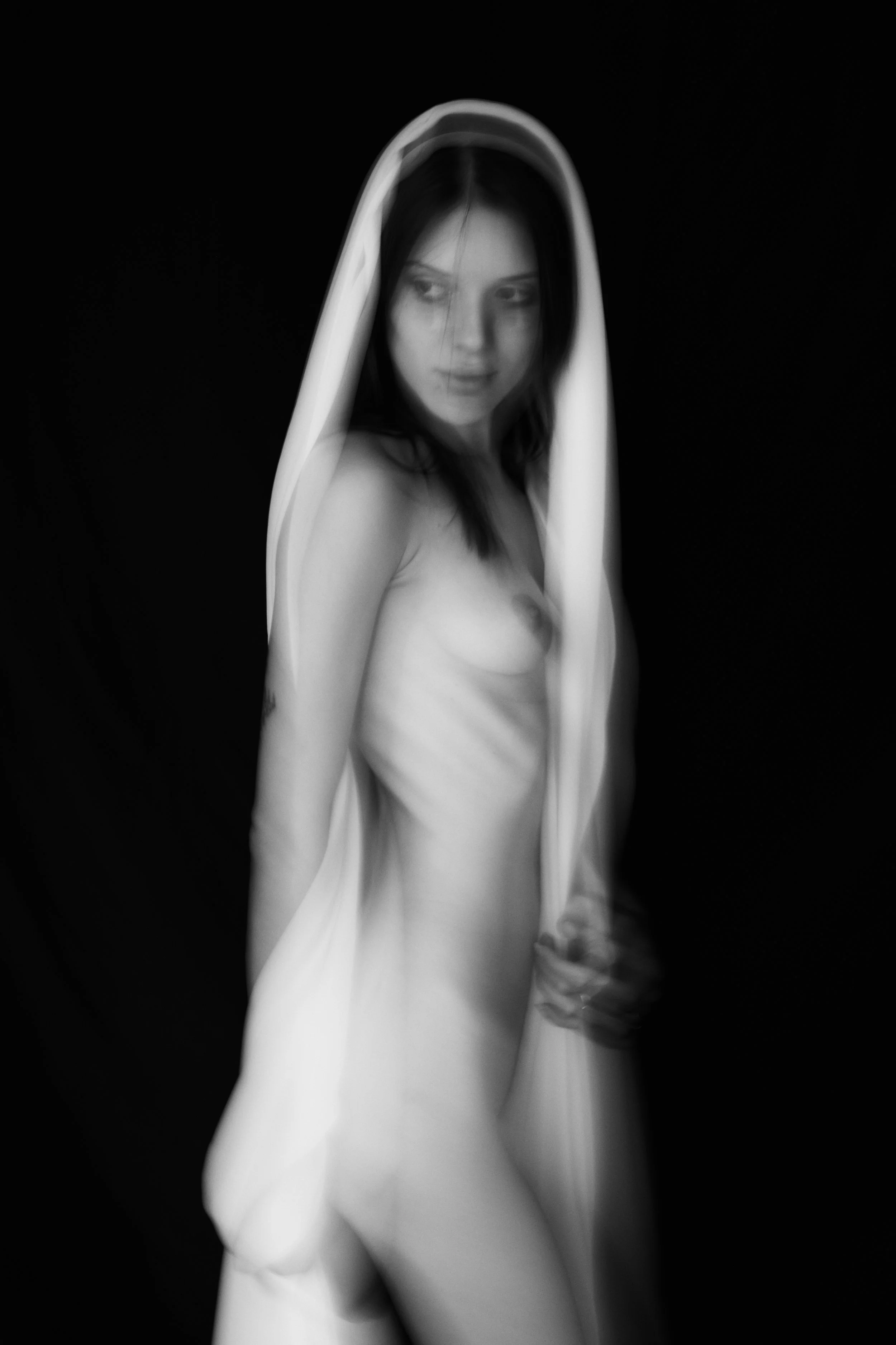 Schwarz-weiße Fotografie einer nackten Frau, die einen weißen Schleier oder Stoff über dem Kopf trägt, vor dunklem Hintergrund, mit nachdenklichem Ausdruck.