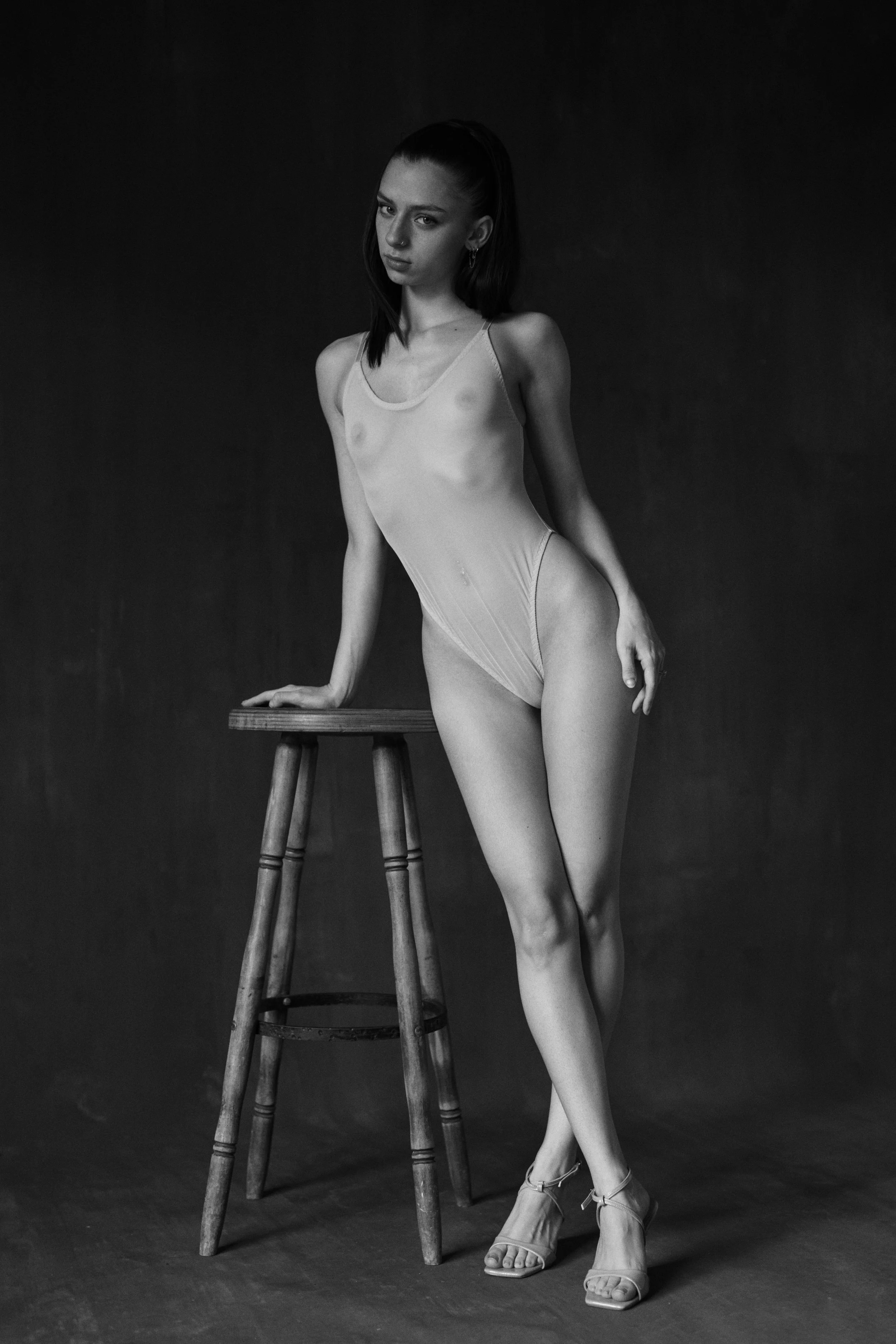Schwarze und weiße Fotografie eines jungen Models in einem transparenten Bodysuit, das auf einem Stuhl lehnt und hochhackige Schuhe trägt.