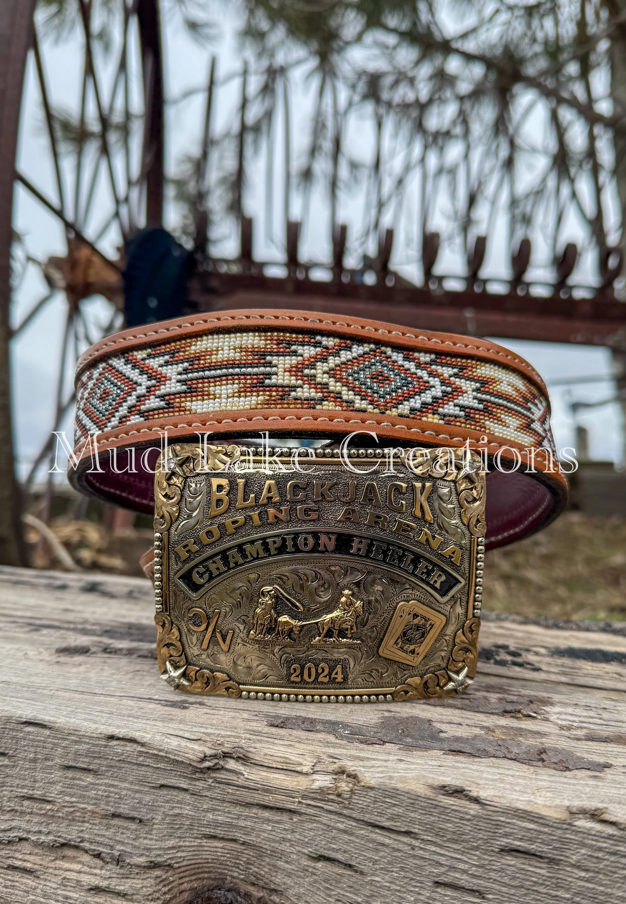Close-up of a large, ornate championship belt with a leather strap and intricate gold and silver detailing, including the words 'Blackjack Roping Arena Champion Heeler 2024', placed on a weathered wooden surface outdoors with a metal fence and cloudy