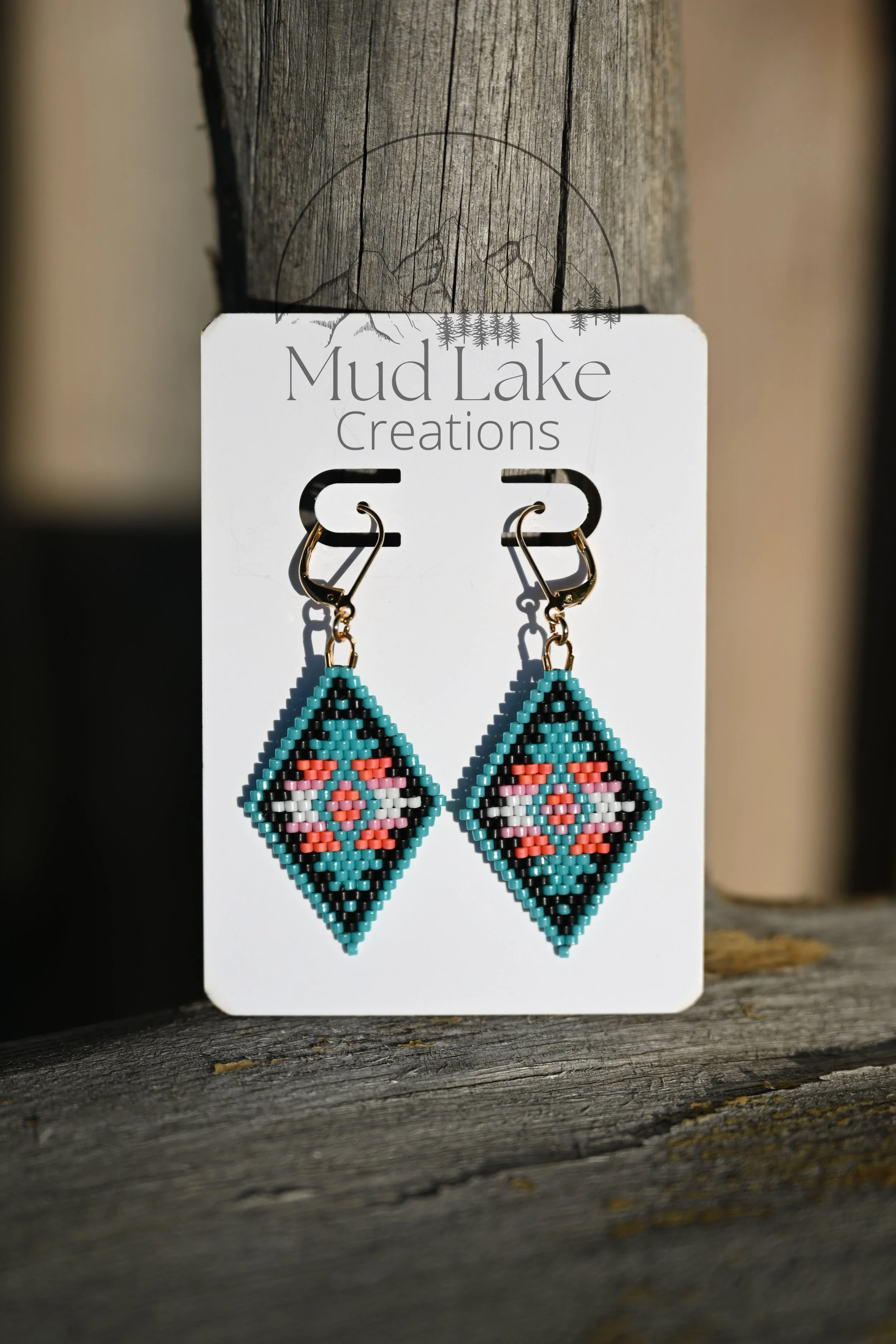 Colorful beaded earrings hanging from a display card with 'Mud Lake Creations' written on it, placed on a weathered wooden surface.