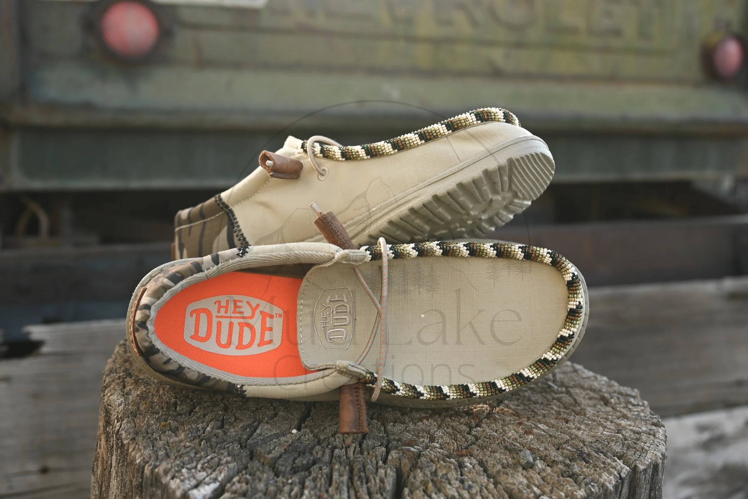 A pair of hiking shoes resting on a tree stump. The shoes are beige with black and orange accents, and have beaded laces. The interior of the shoe displays the brand 'Hey Dude'.