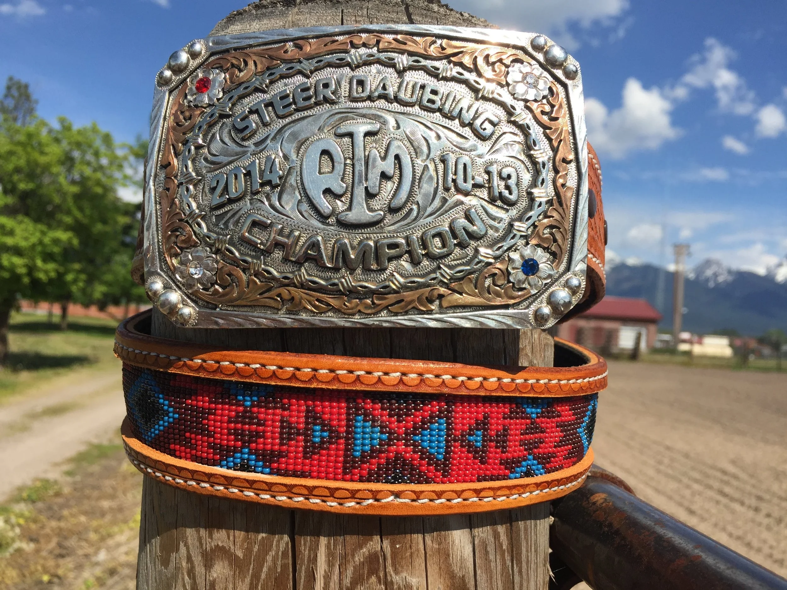 A silver decorative belt buckle with engraved text and design, mounted on a ranching or Western-style belt, against a backdrop of trees, mountains, and a blue sky with clouds.