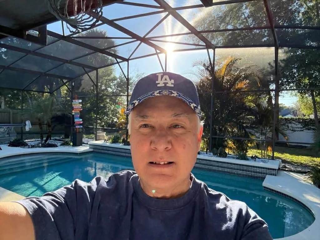 A man taking a selfie outdoors by a backyard pool with a screened enclosure, sun shining through, surrounded by trees and plants.