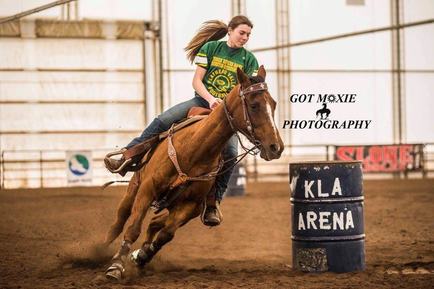 A female horse rider wearing a green jersey riding a brown horse around a barrel at an indoor arena.