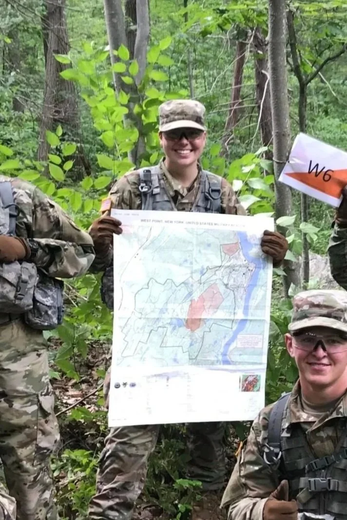 A group of diverse soldiers in camouflage uniforms and tactical gear standing in a forest. They are holding a map, a flag, and smiling, indicating a sense of achievement or teamwork.