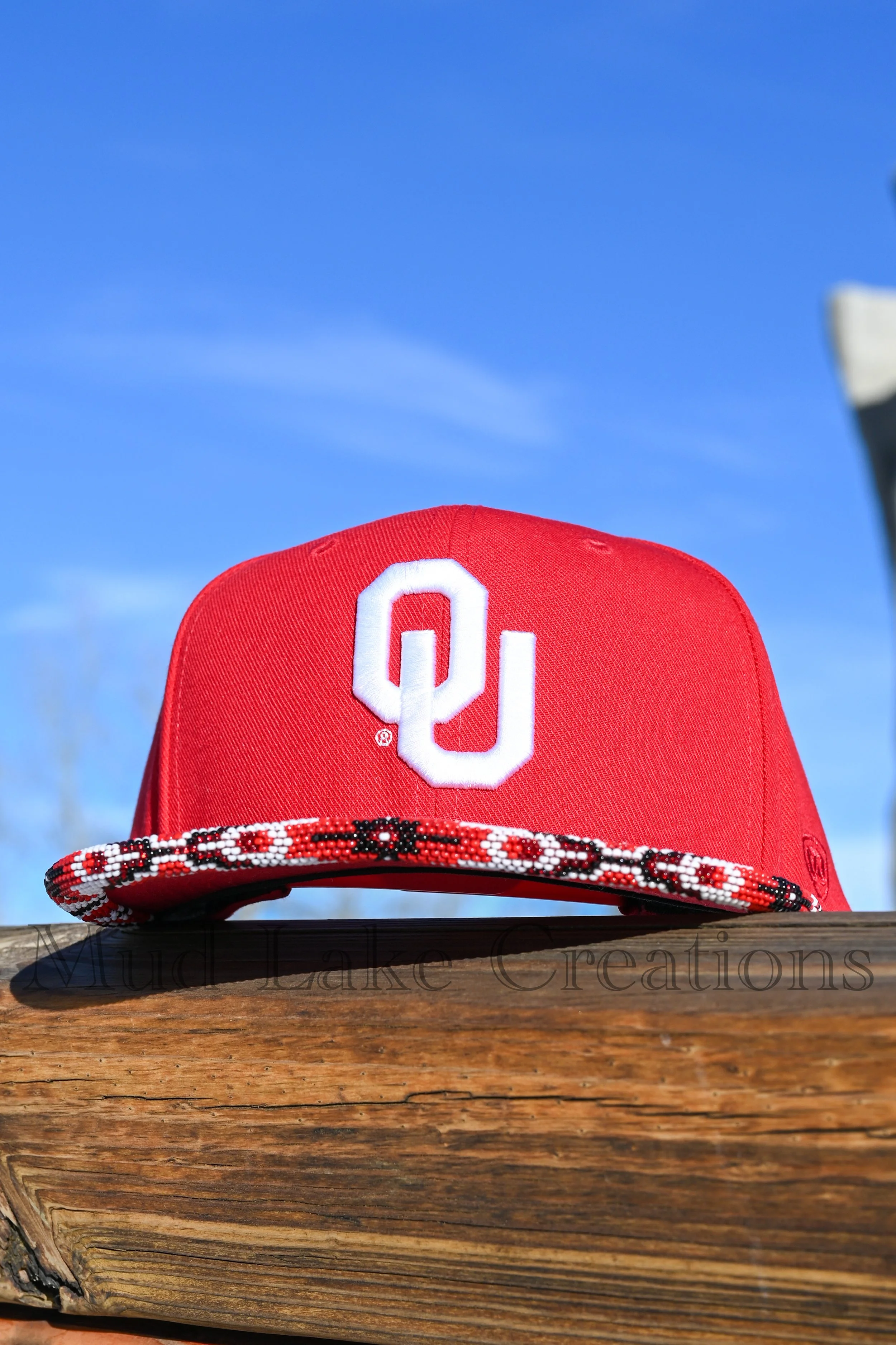 A red Oklahoma Sooners baseball cap with a white embroidered letter 'O' on the front, sitting on a wooden surface against a clear blue sky.