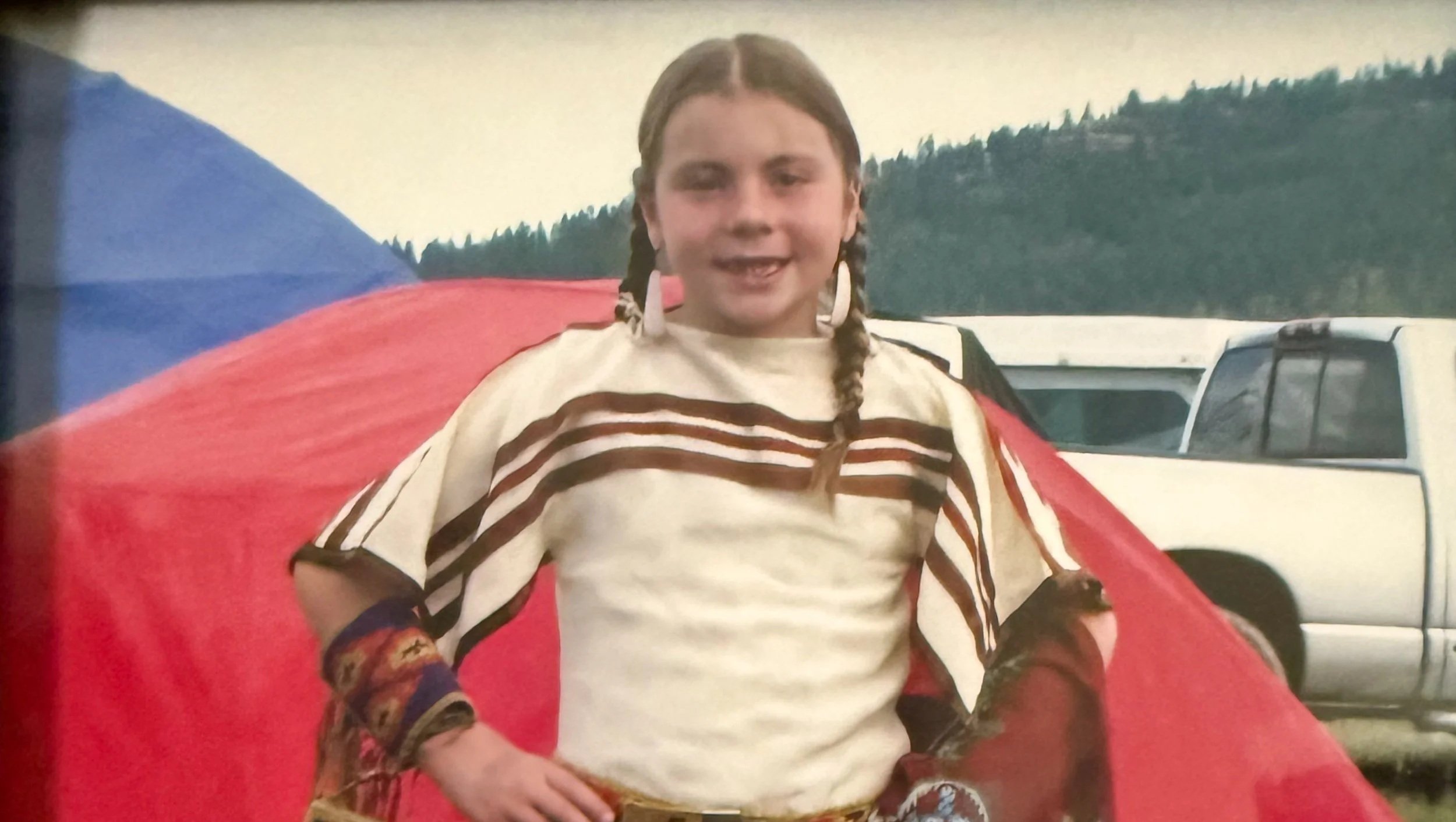 A young indigenous girl with braided hair smiling, sitting in front of a red tent, with a white pickup truck and a forested hillside in the background.