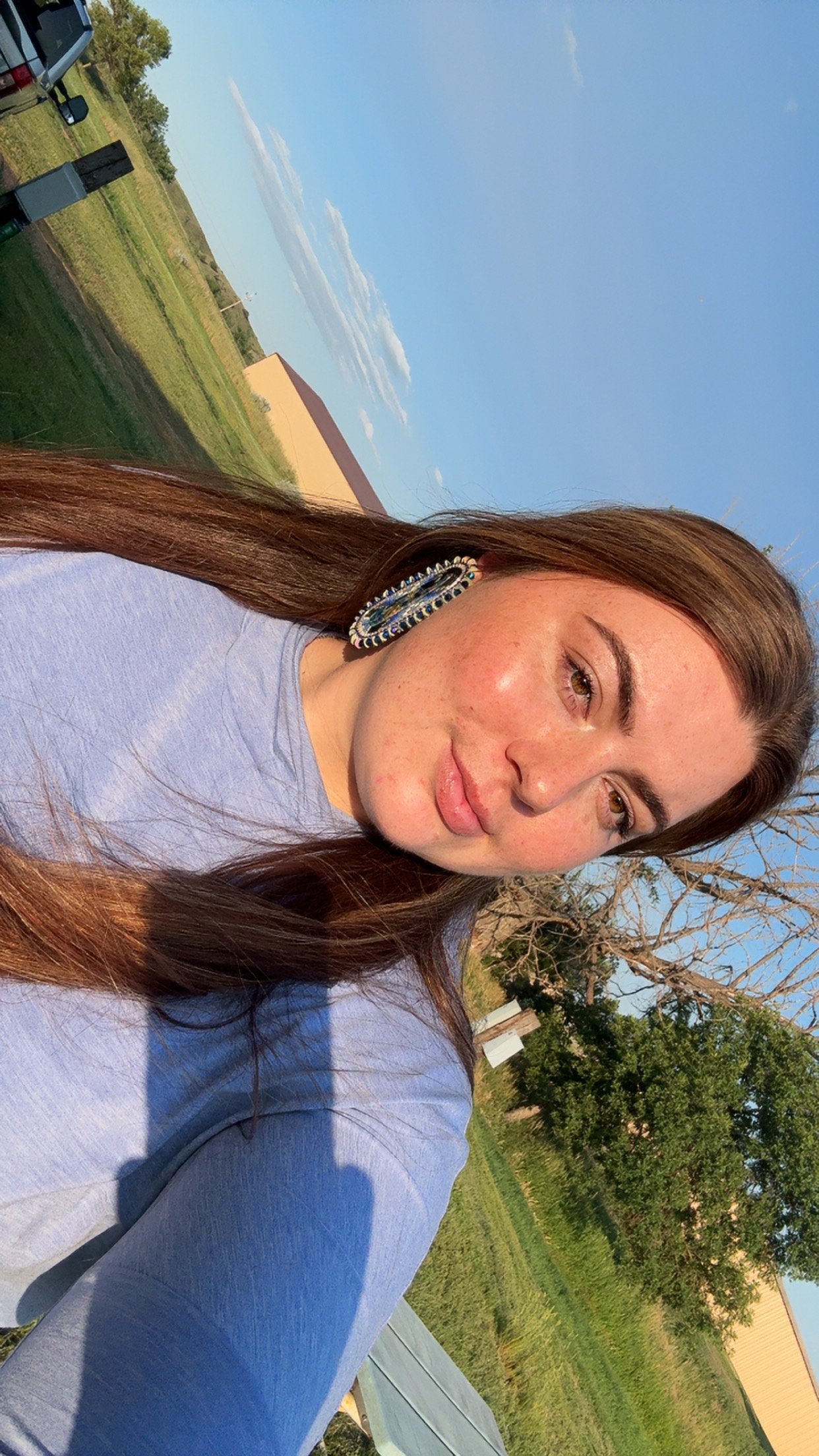 A young woman with long brown hair and hoop earrings taking a selfie outdoors on a sunny day, with green grass, trees, and a building in the background.