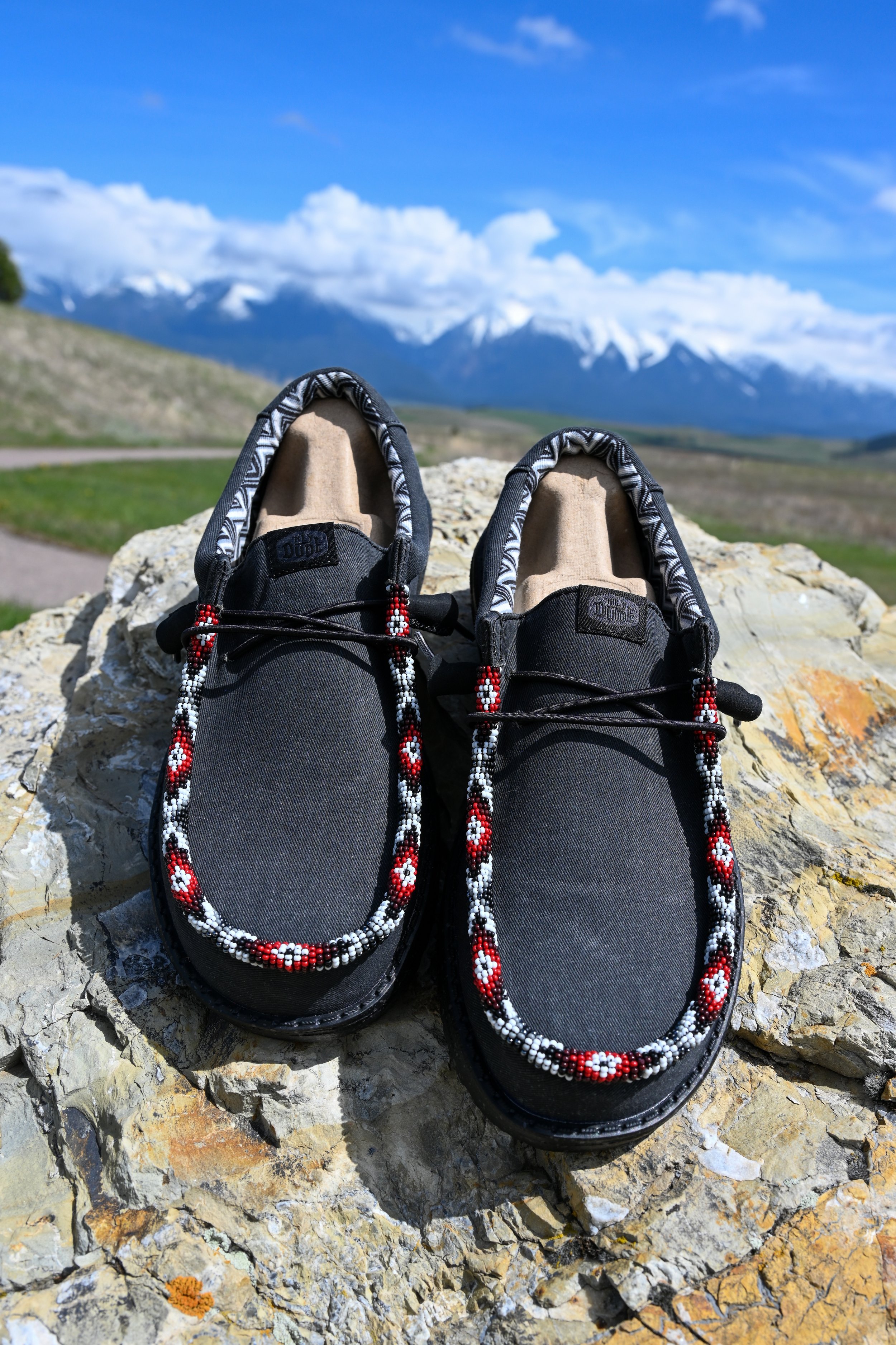 Pair of black shoes with red, white, and black beaded jewelry resting on a rocks surface outdoors with green fields, snow-capped mountains, and a blue sky with scattered clouds in the background.