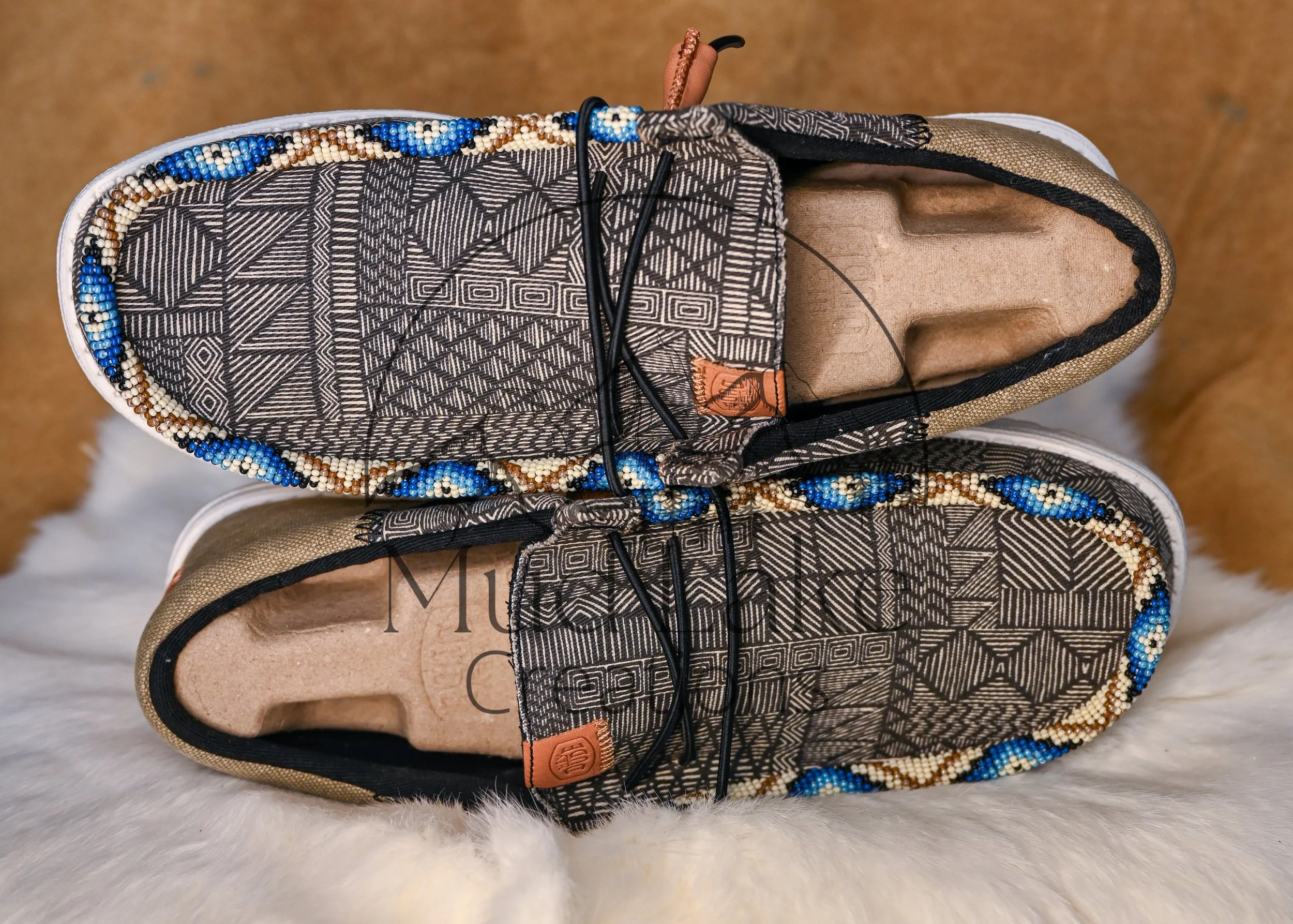A pair of Tribal Beaded Sneakers with detailed black and white geometric patterns, beaded trim, and linen interior, placed on a soft white furry surface.