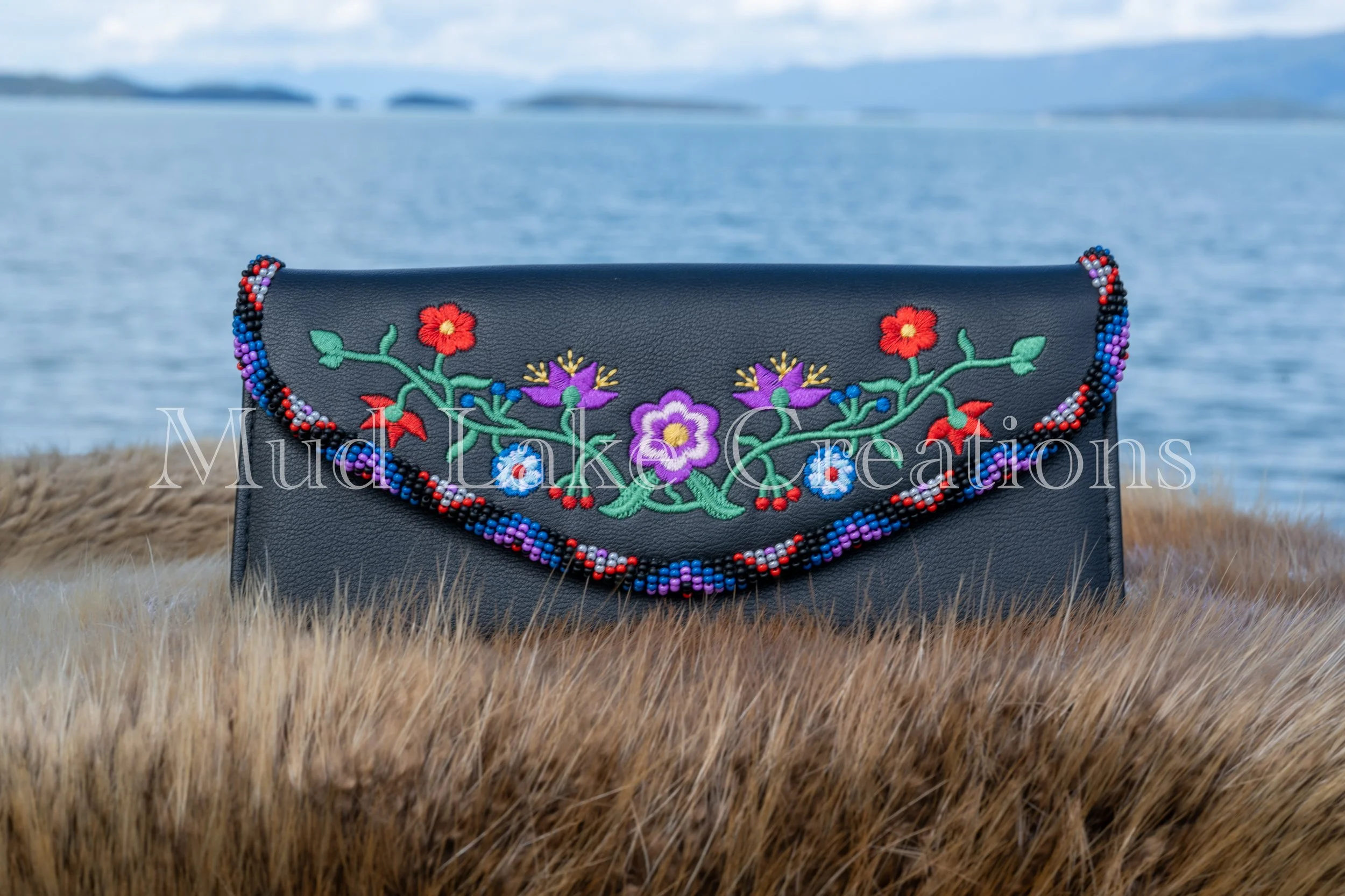 Black purse with colorful embroidered floral design and beaded trim, placed on tall grass near water with islands in the distance and cloudy sky