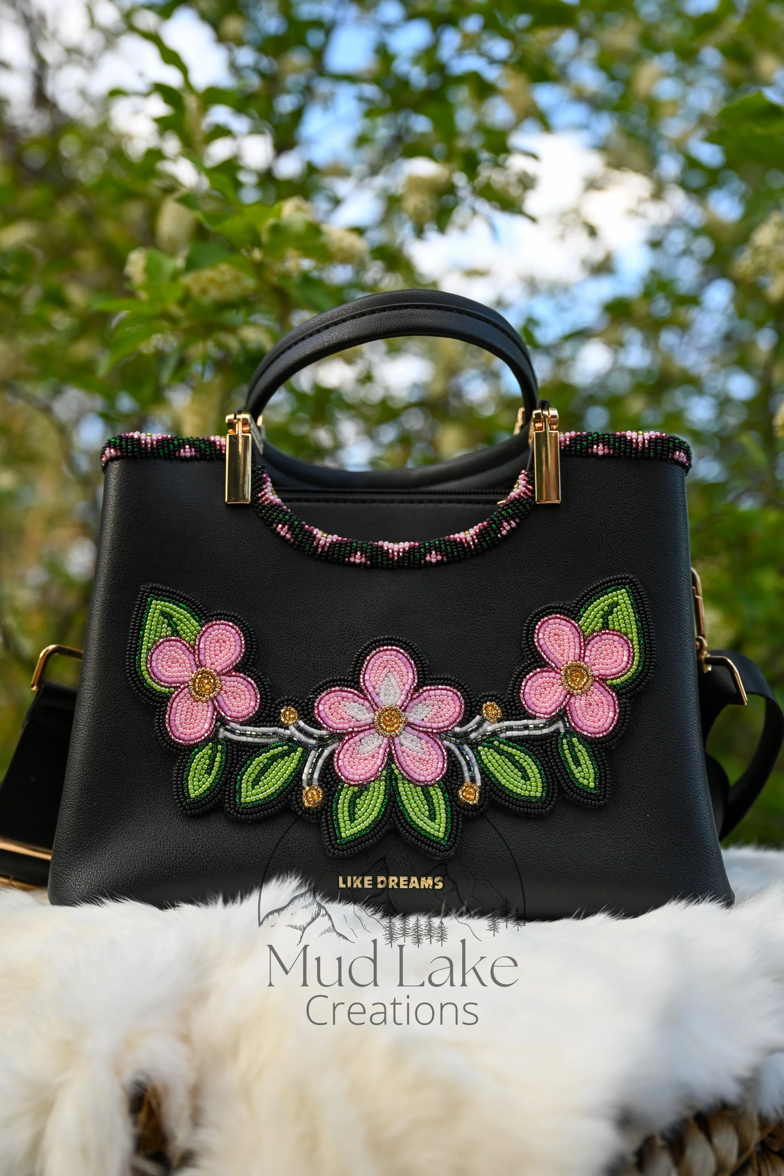Black handbag with colorful floral bead beadwork, gold accents, and the text 'Mud Lake Creations' on it, displayed outdoors against a background of green leaves and blue sky.