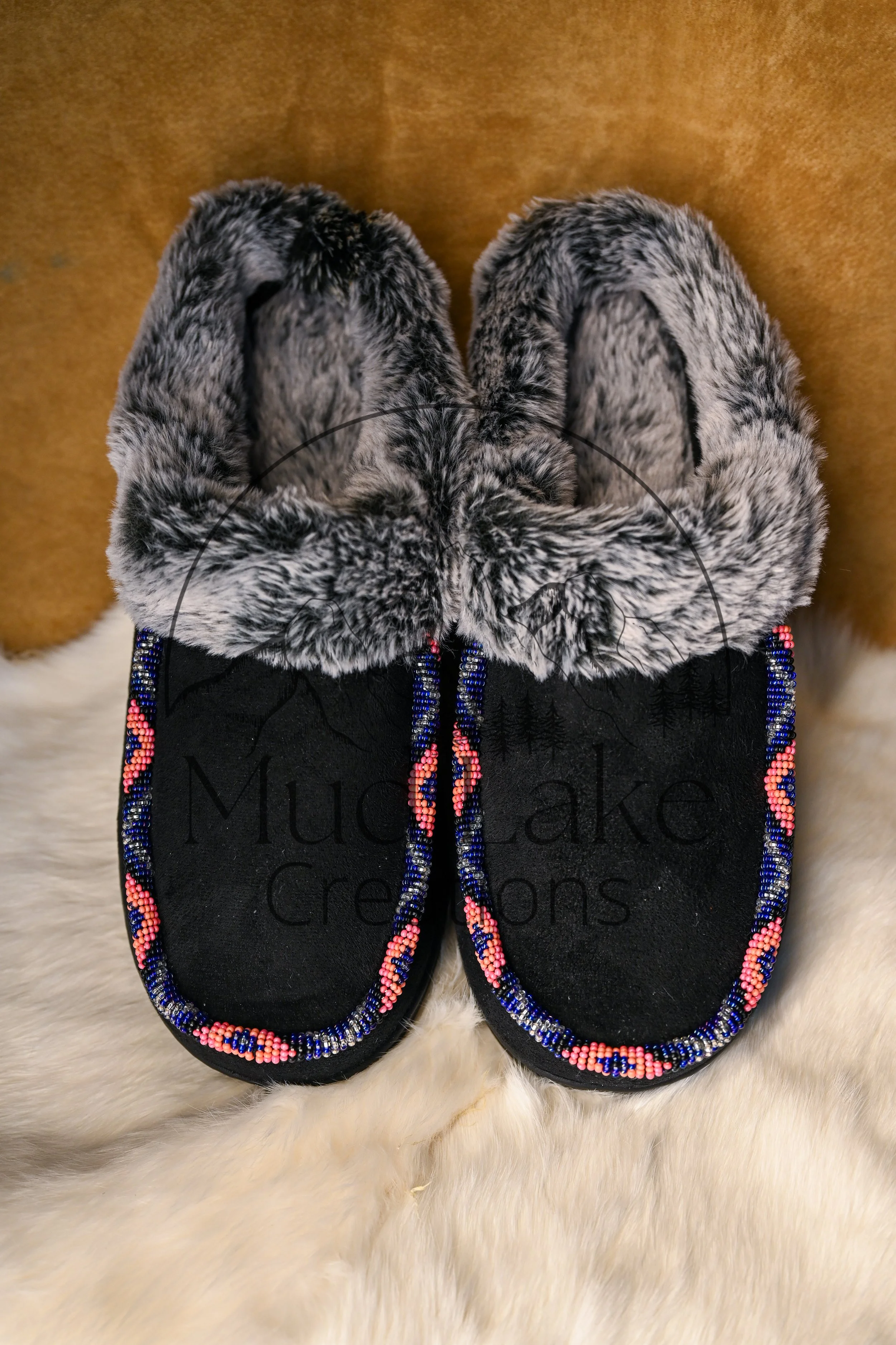 Pair of black moccasins with colorful beaded decorations around the edges, placed on a cream faux fur surface. The shoes have faux fur lining and beaded strings attached, with one shoe slightly overlapping the other.