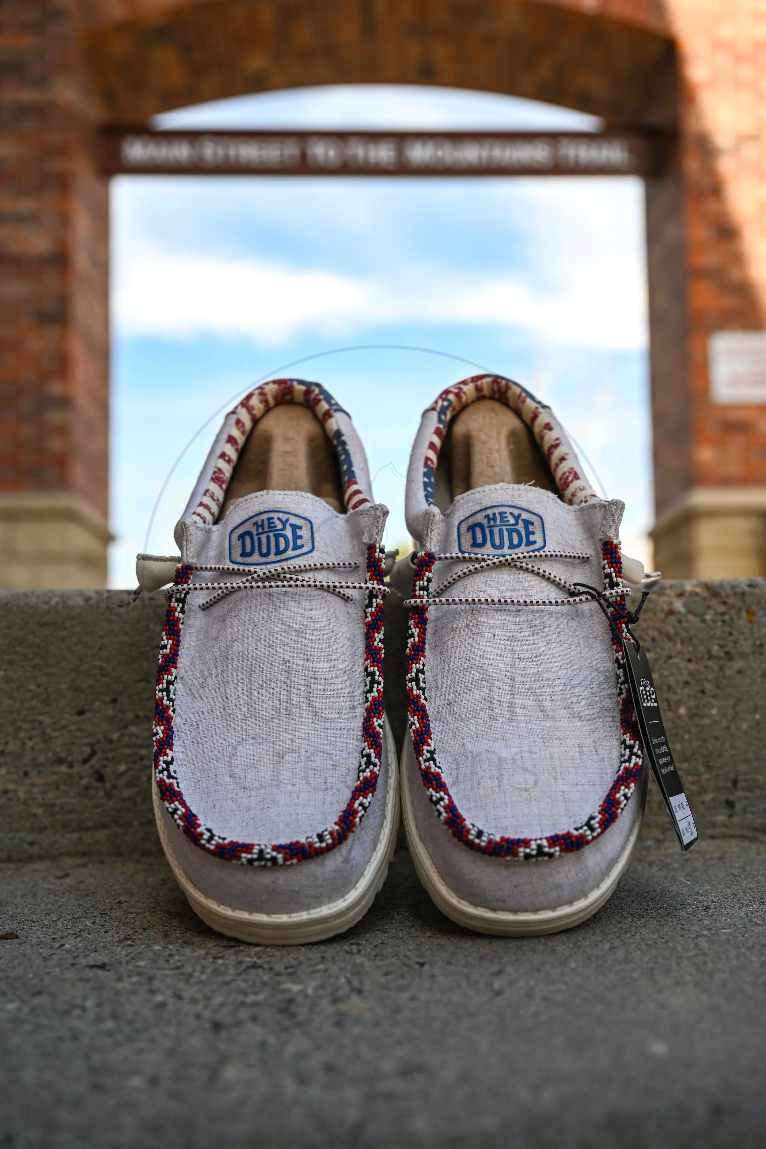 A pair of beaded Hey Dudes with colorful beaded patterns and blue 'Hey Dude' logos on the tongues, placed outdoors on a concrete surface with a brick archway and a cloudy sky in the background.