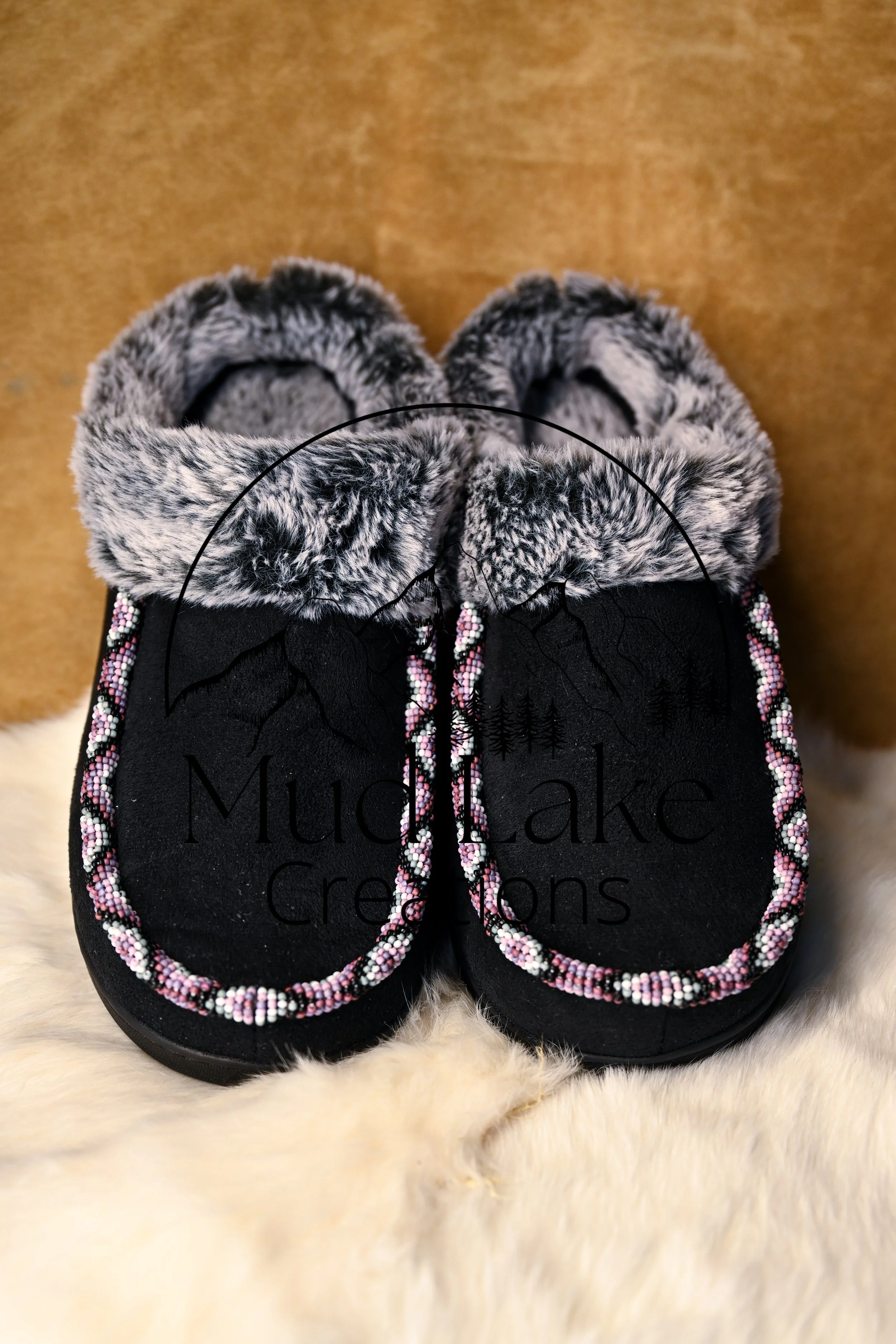 Pair of black moccasins with pink and white beadwork and faux fur trim, displayed on a white furry surface.