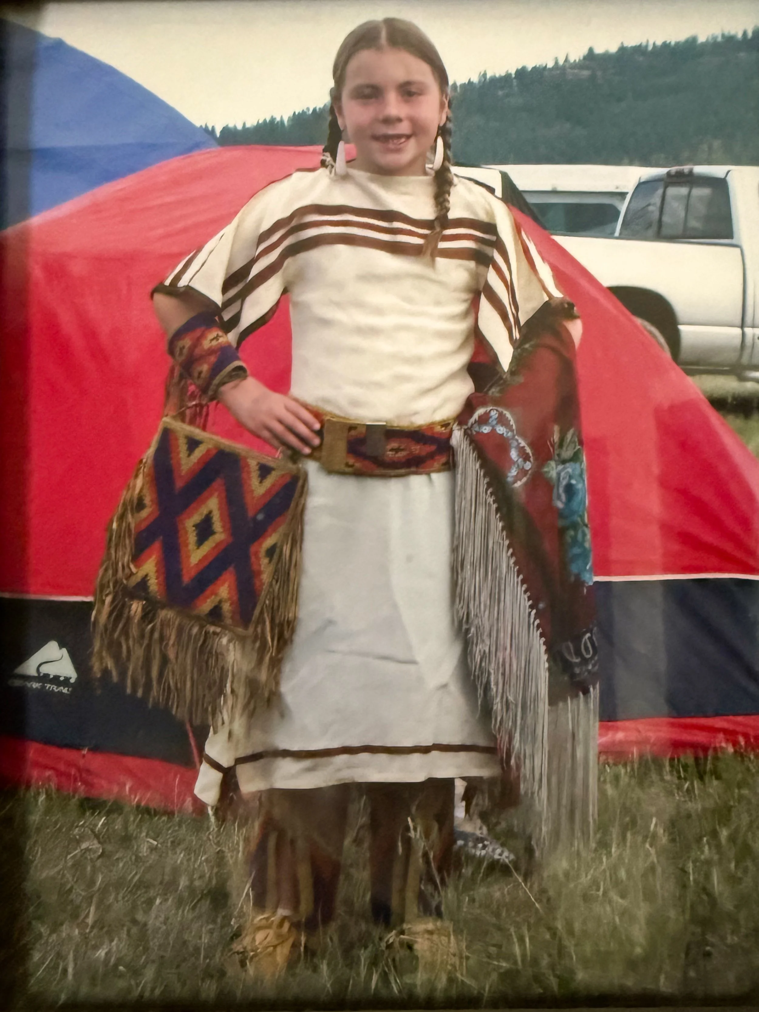 A young girl with braided hair wearing a white dress and fringed shawl, standing outside near a red tent and white trucks, with forested hills in the background.