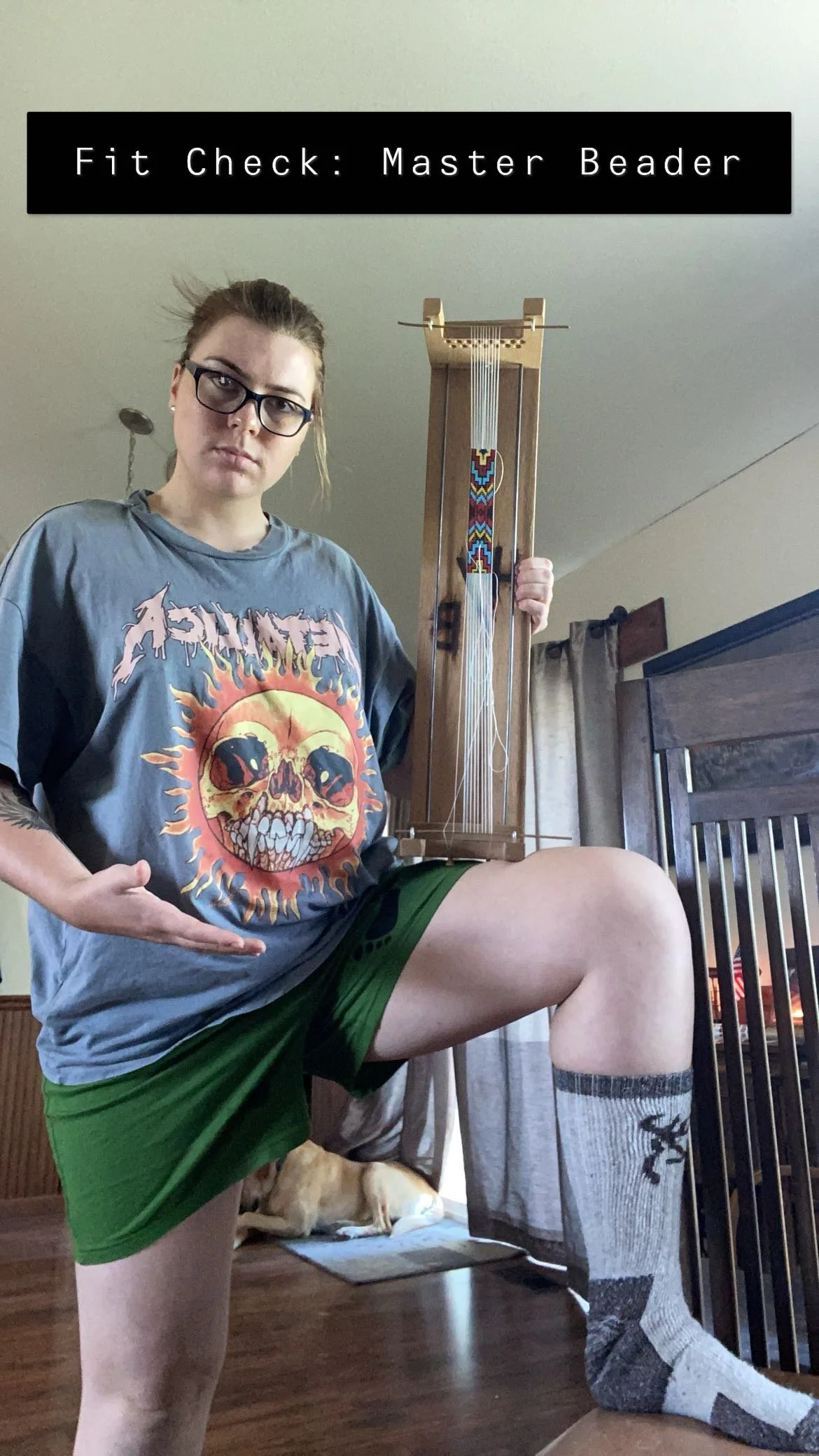 A woman wearing a Thrasher T-shirt, knee-high sock, and shorts poses with one knee up on a chair, holding a loom, with a dog lying underneath. She has glasses and short hair.