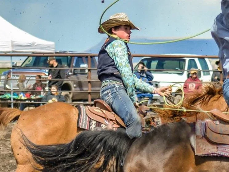 Young boy riding a brown horse at a rodeo event, wearing a cowboy hat, plaid jeans, and a patterned shirt, with spectators and vehicles in the background.