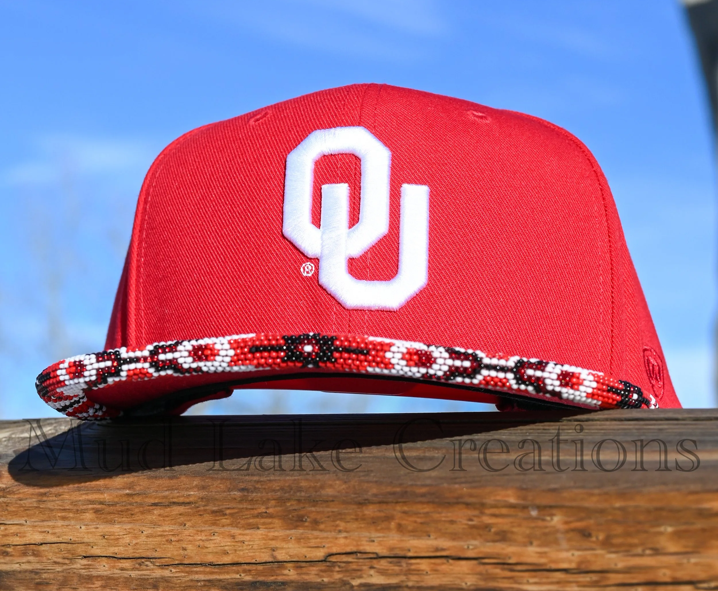 Close-up of a red baseball cap with the University of Oklahoma logo, sitting on a wooden surface with a peyote stitched beaded brim, against a blue-sky background.