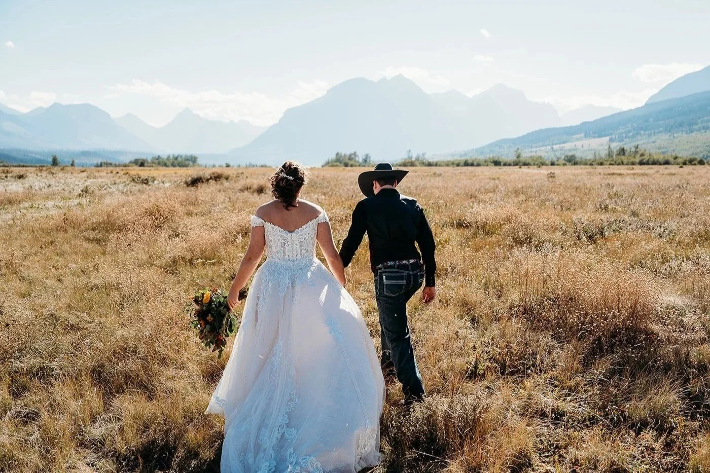 Couple in wedding attire walking through a field with mountains in the background, the woman is wearing a white wedding dress and holding a bouquet, the man is wearing a black cowboy hat and dark clothing, with a western beaded belt.