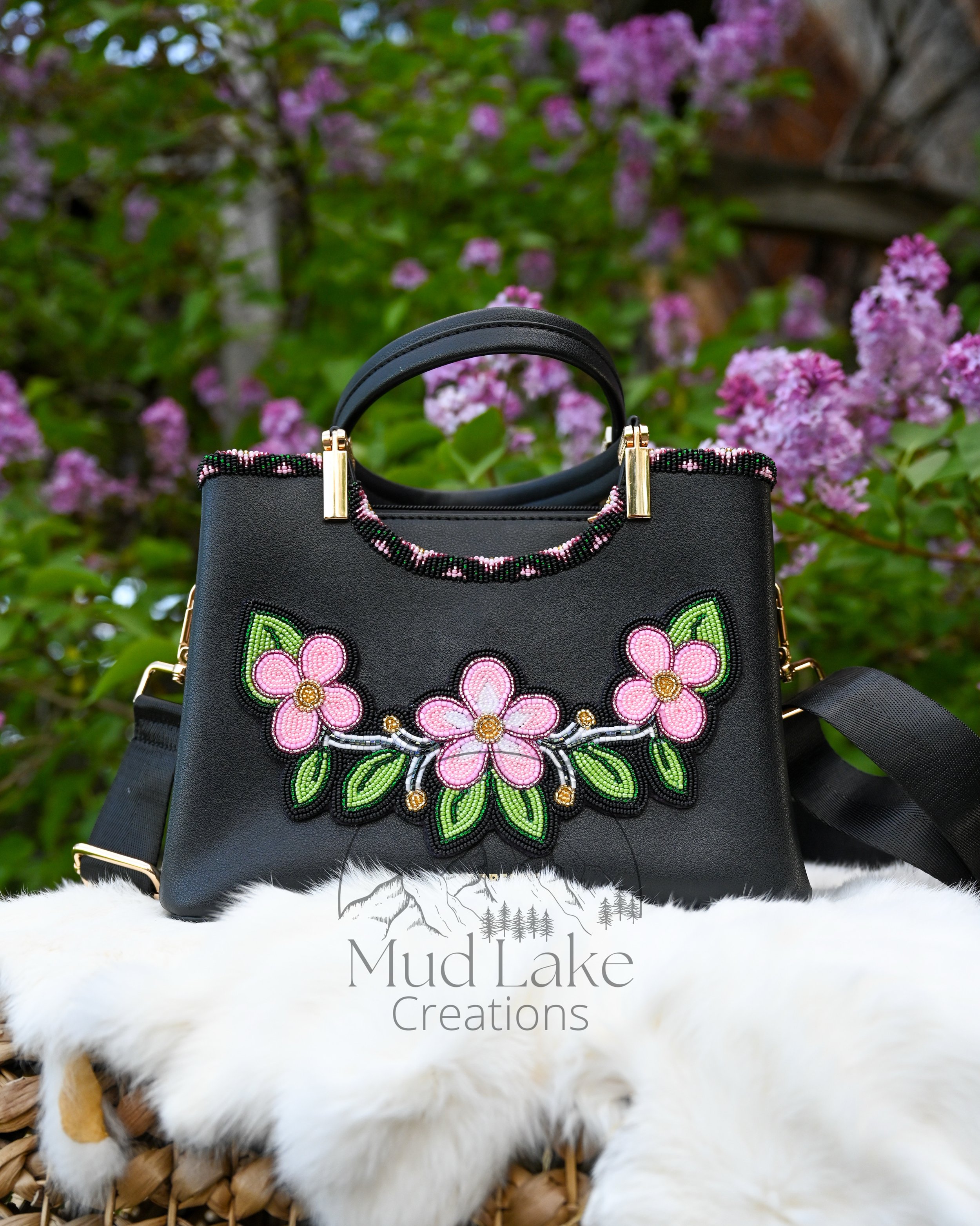 A black handbag with floral embroidery and beaded accents, placed on a white furry surface with a basket and pink flowering bushes in the background.