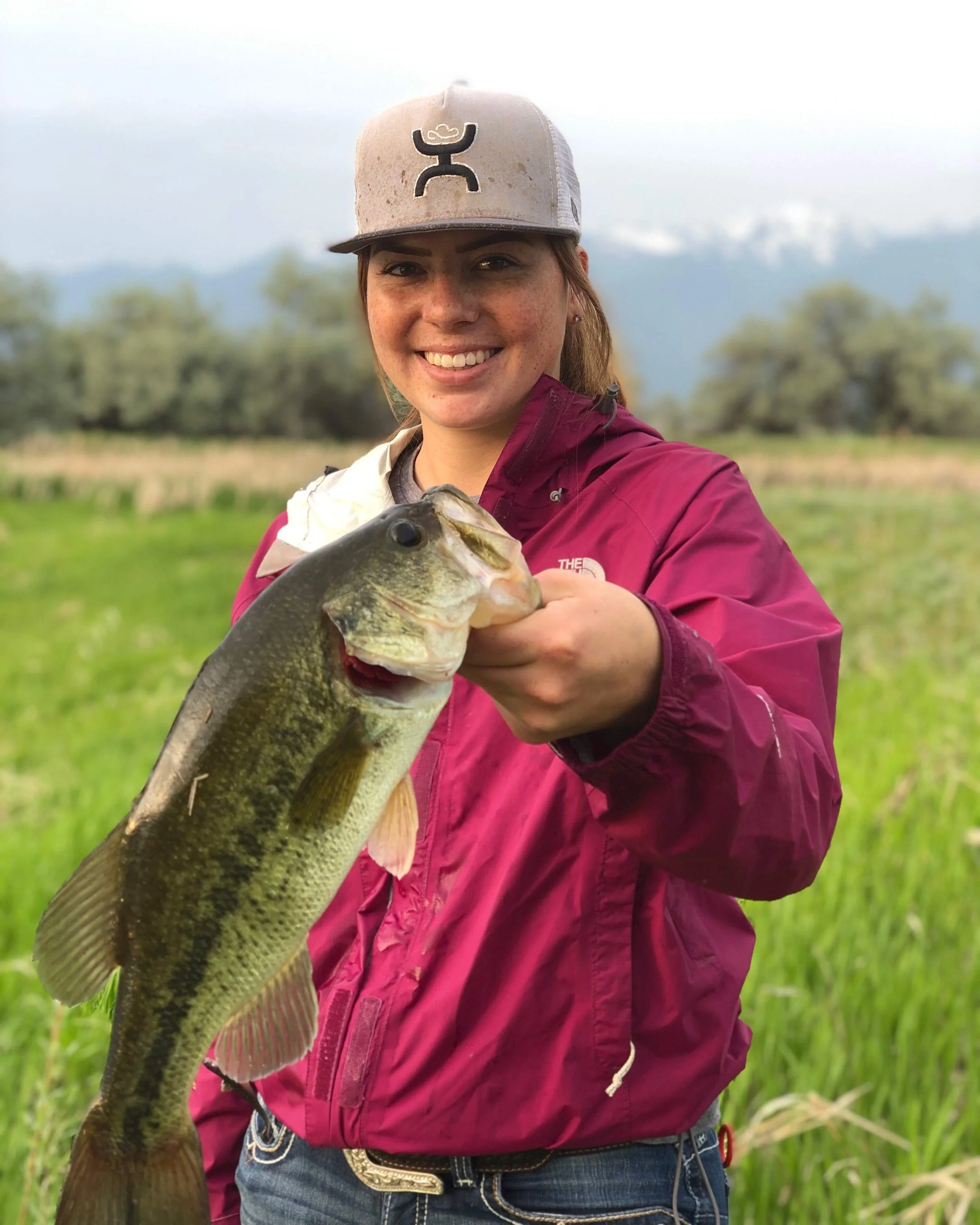 Smiling woman holding a large fish outdoors with green grass and trees in the background, wearing a beige cap with a black logo, a maroon jacket, and jeans.