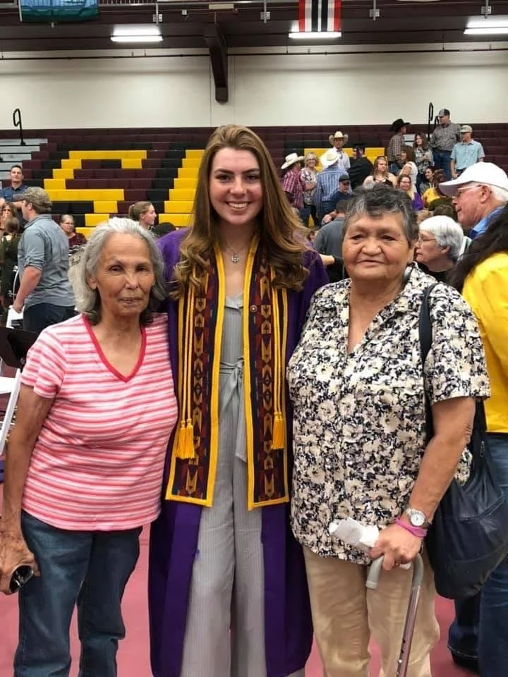 Young woman in graduation gown and stole standing with two older women at a graduation ceremony in a gymnasium, with other attendees in the background.
