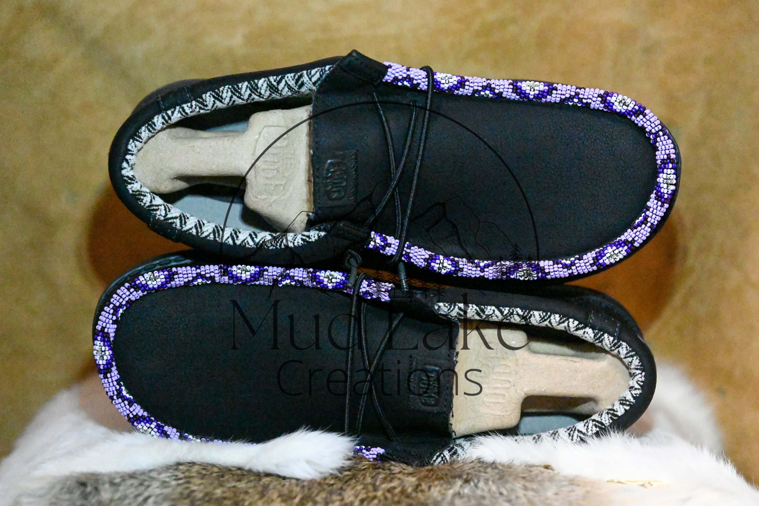 Close-up of a black sneaker with purple, white, and black beaded details along the edge, placed on a furry surface with a tan background.