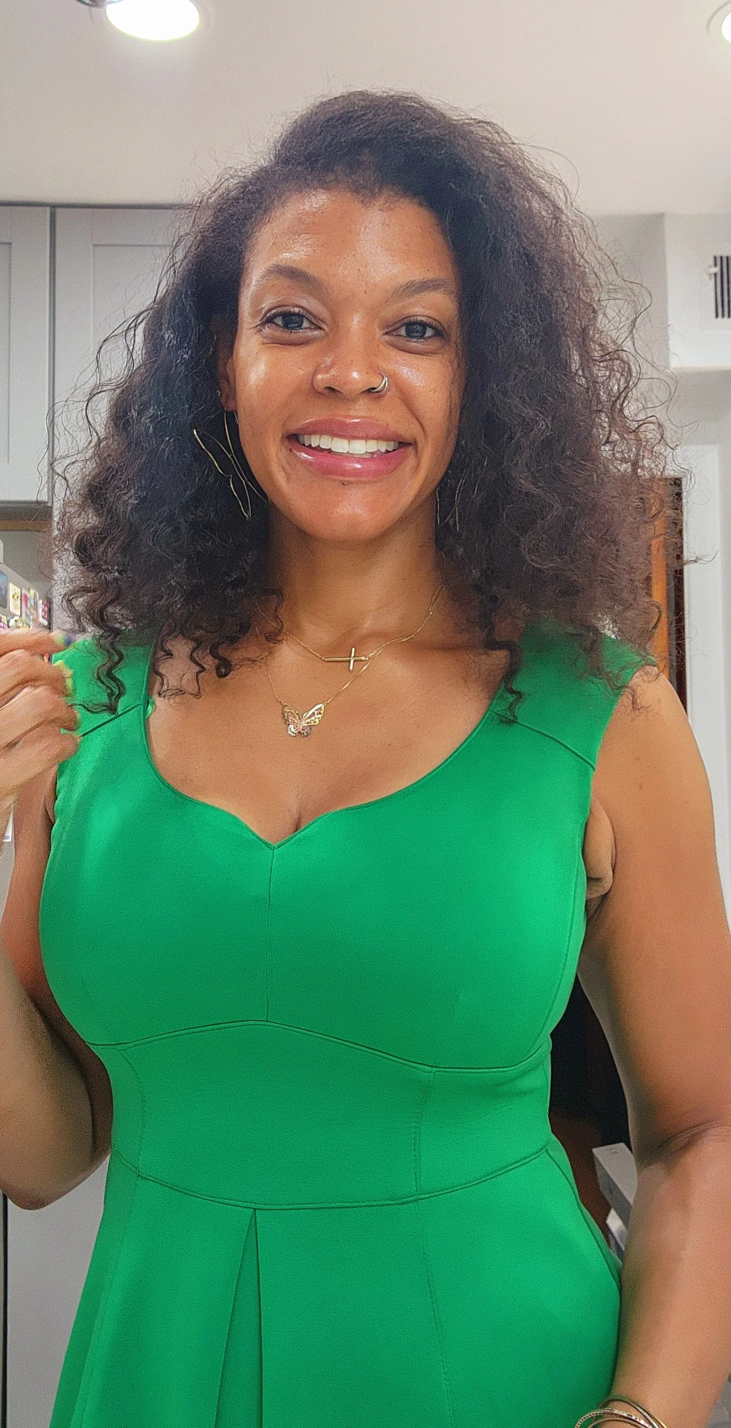 A woman with curly black hair wearing a green sleeveless dress, smiling indoors, accessorized with layered necklaces and earrings.