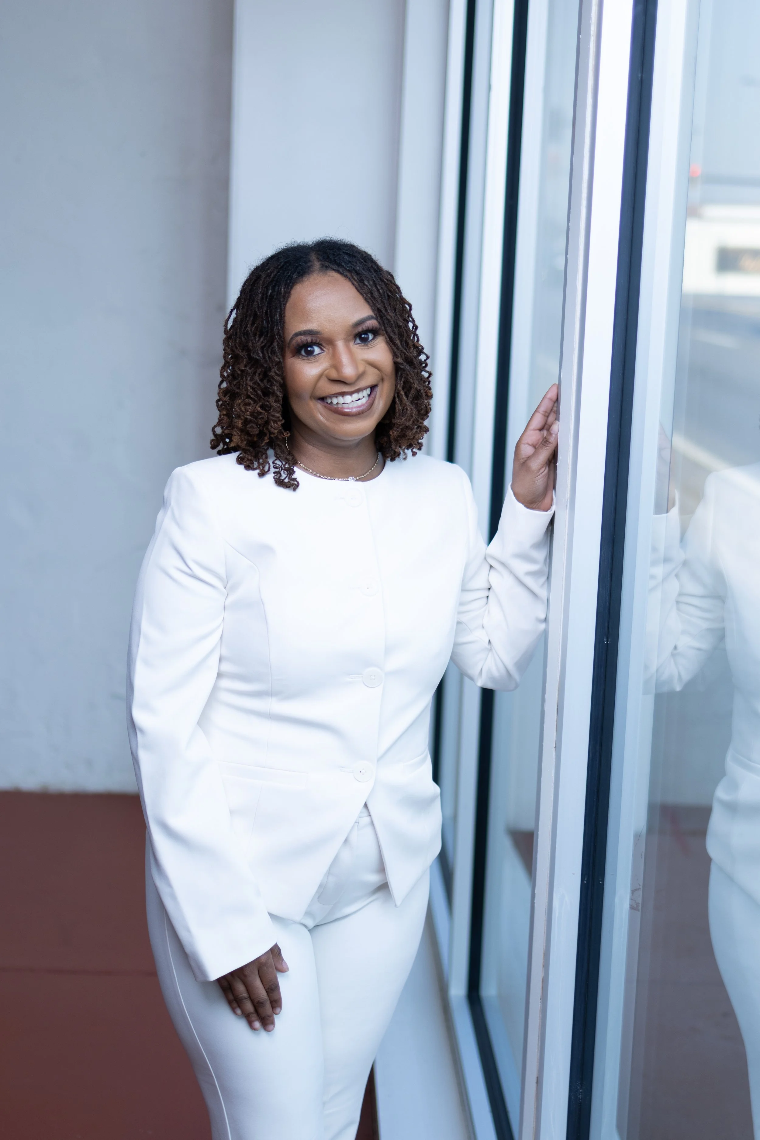 A woman dressed in a white suit and white pants smiling and standing beside a large window in an indoor setting.