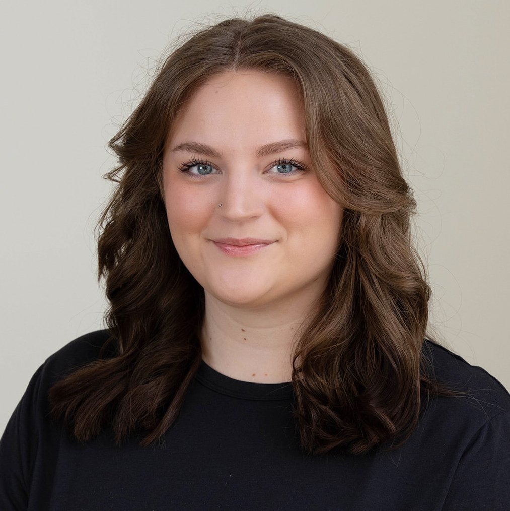 A trainer at Infinity Fitness in Reno with long curly brown hair, wearing glasses and a sleeveless black top, standing in front of a wooden wall, smiling with her hands on her hips.