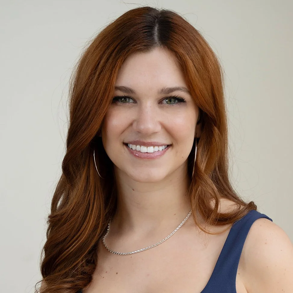 A trainer at Infinity Fitness in Reno with shoulder-length auburn hair, wearing a black workout sports bra and matching high-waisted leggings, standing in front of a wooden wall, smiling at the camera.