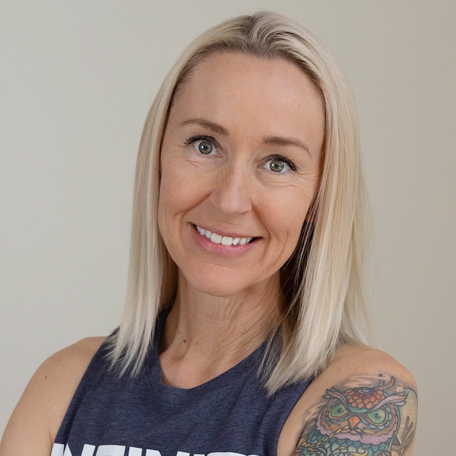 A trainer with long blonde hair with blue tips, smiling and crossing her arms, standing against a wooden wall. She is wearing a sleeveless gray shirt with the word 'INFINITY' 