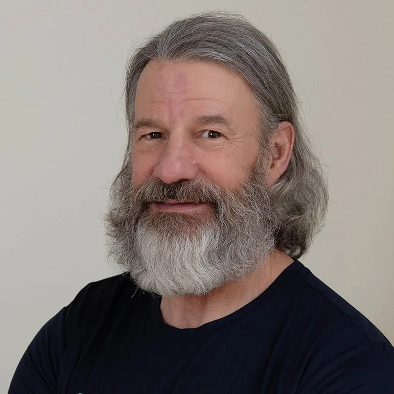 A trainer at Infinity Fitness in Reno with a beard smiling, wearing a blue t-shirt that says 'INFINITY FITNESS STUDIOS', standing against a wooden wall background.