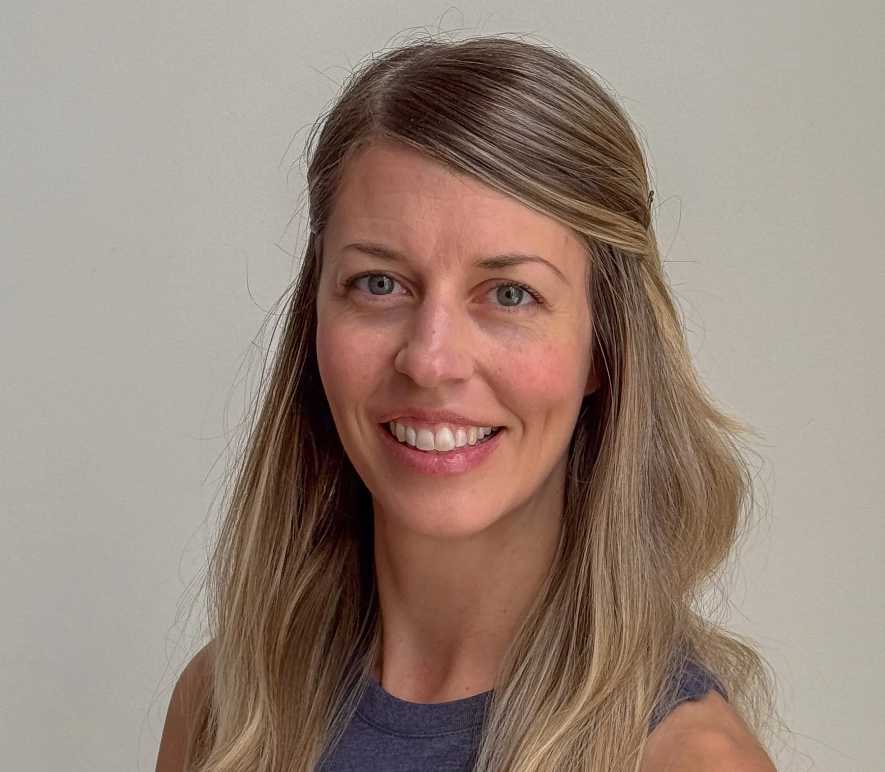 A trainer at Infinity Fitness in Reno with long blonde hair and blue eyes, smiling, wearing a black top, standing against a wooden wall background.