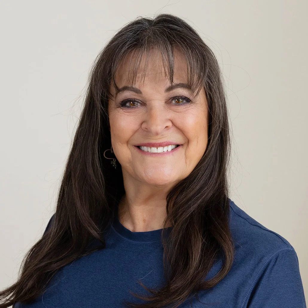 Smiling trainer at Infinity Fitness in Reno with dark hair, wearing a black T-shirt that reads "INFINITY FITNESS STUDIOS," standing against a wooden wall.