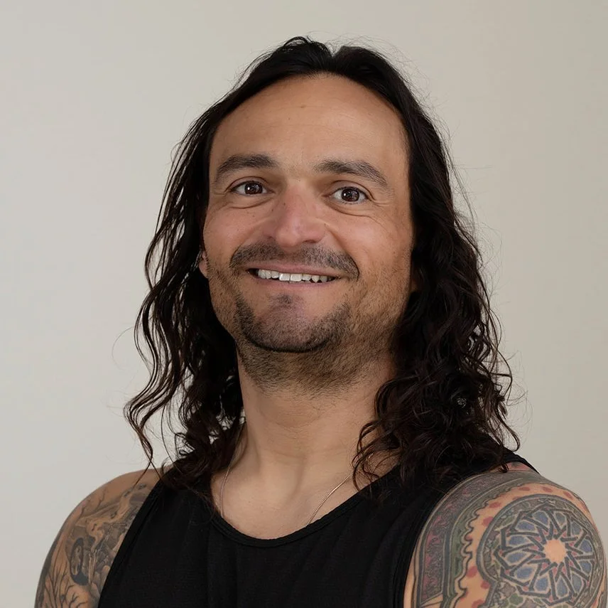 A trainer at Infinity Fitness in Reno with long dark hair and a beard, smiling with arms crossed, wearing a black sleeveless shirt, standing in front of a wooden wall.