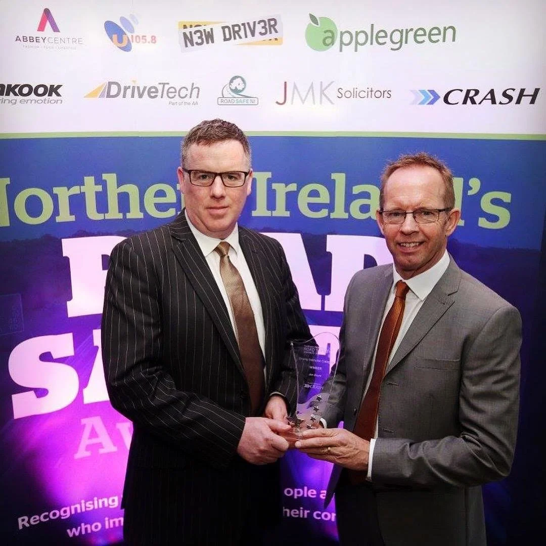 Jim Doyle recieving a glass award at an event with a backdrop displaying logos and text, including 'Northern Ireland's' and 'P... S... A...'.