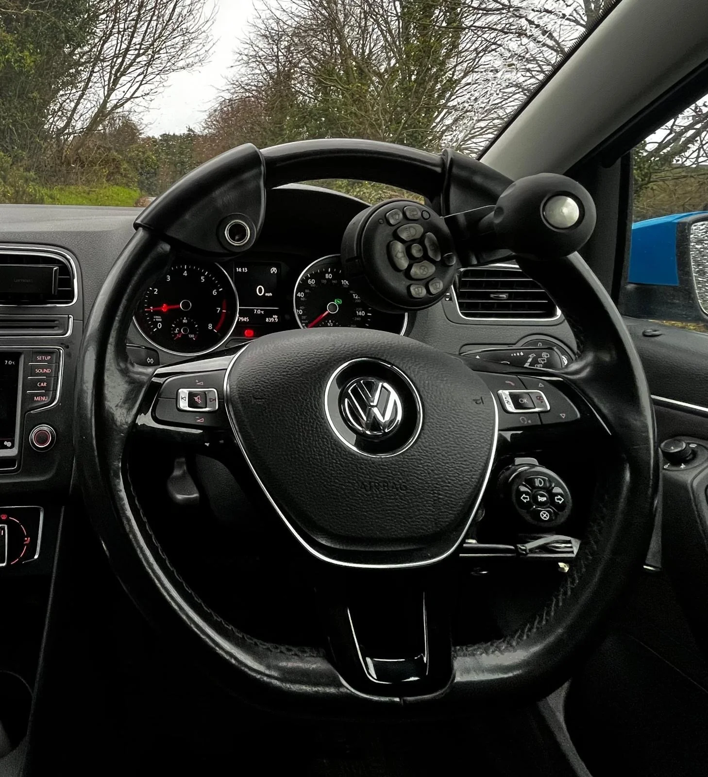 The interior of a Volkswagen car shows a steering wheel with the VW logo, dashboard with speedometer and tachometer, and various controls. Outside scenery with trees and an overcast sky is visible through the windshield.