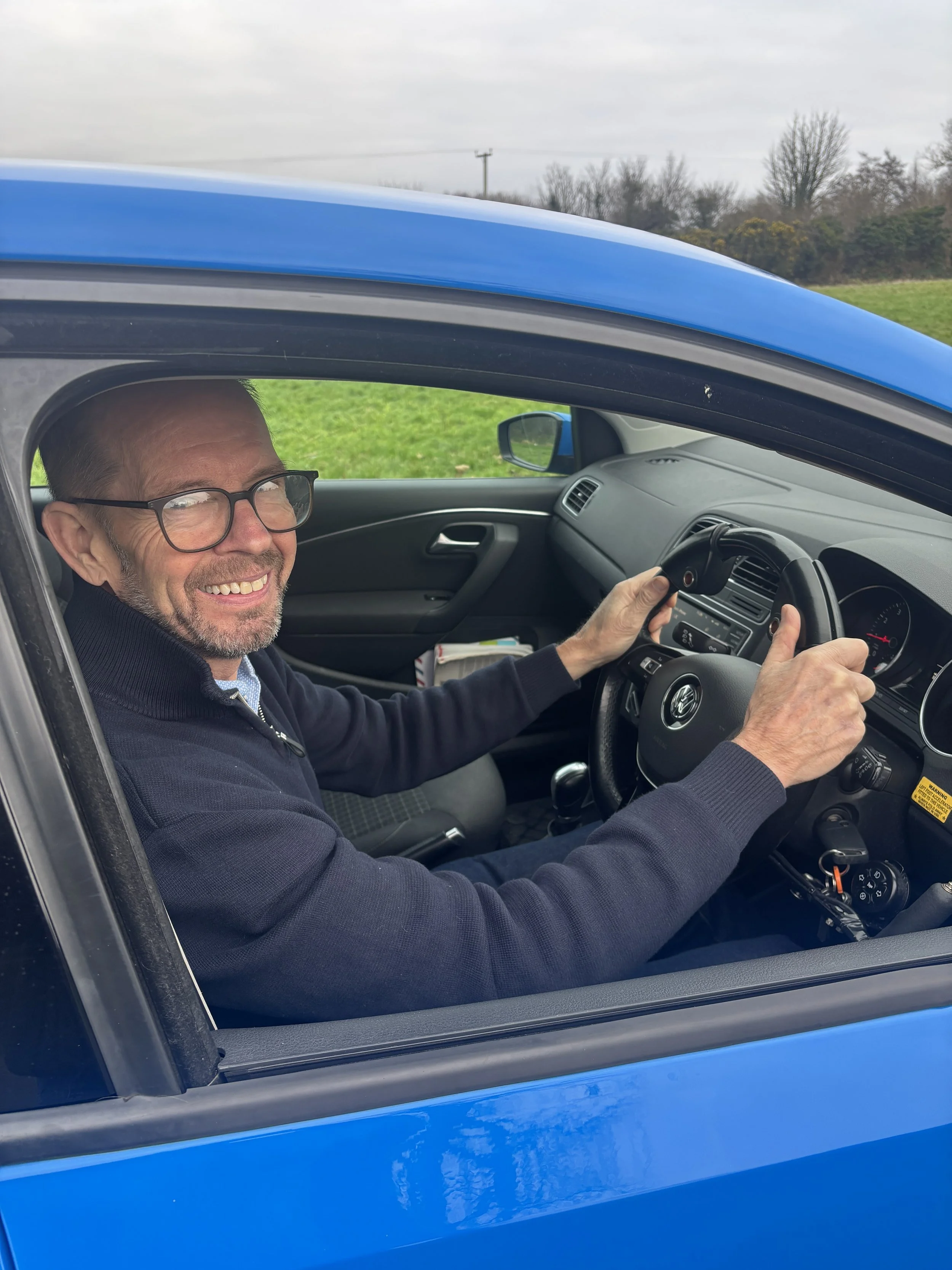 Jim Doyle smiling sat in the driver’s seat of a blue car, holding the steering wheel with both hands, with a rural landscape of grass and trees outside.