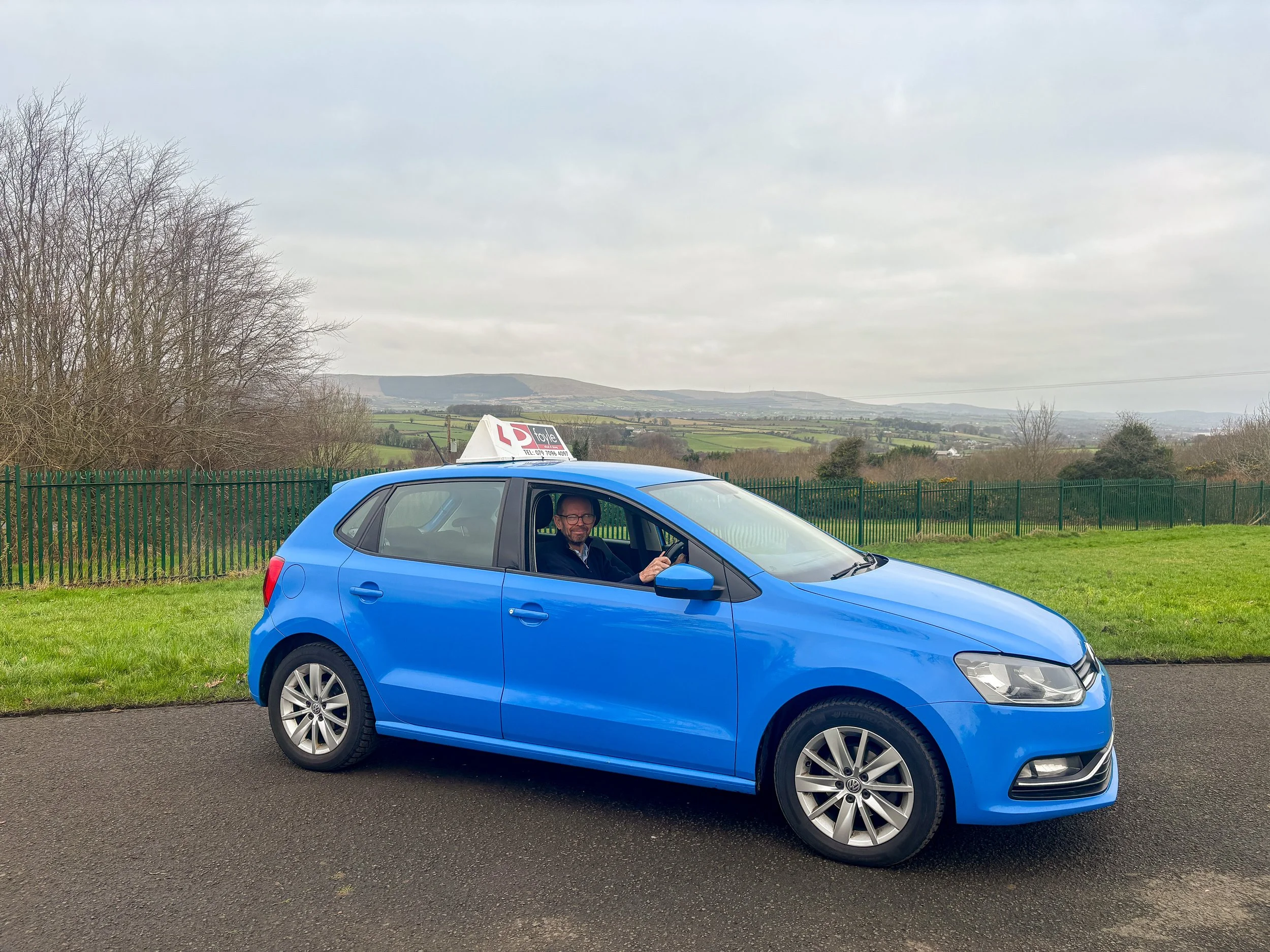 Jim Doyle sitting inside a blue Volkswagen car with the Foyle School of Driving school sign on top, parked on a road near a green fence and grassy area with trees and hills in the background.