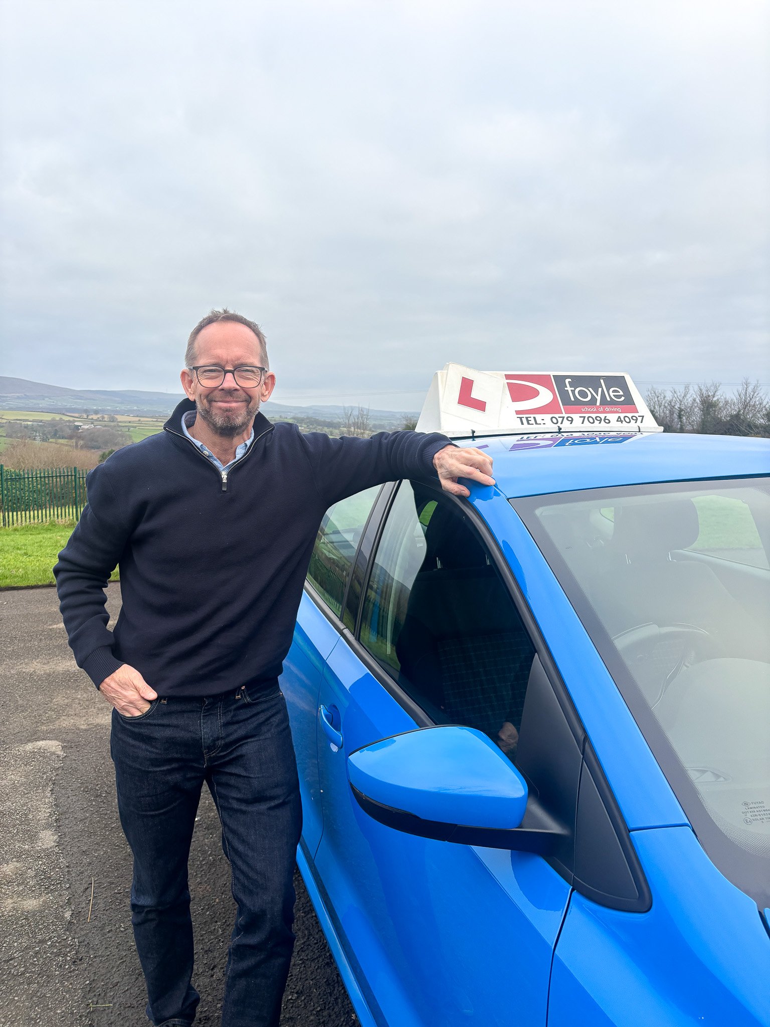 Jim Doyle standing next to the Foyle School of Driving blue car with a learner driver sign on top, outdoors with a cloudy sky and countryside in the background.